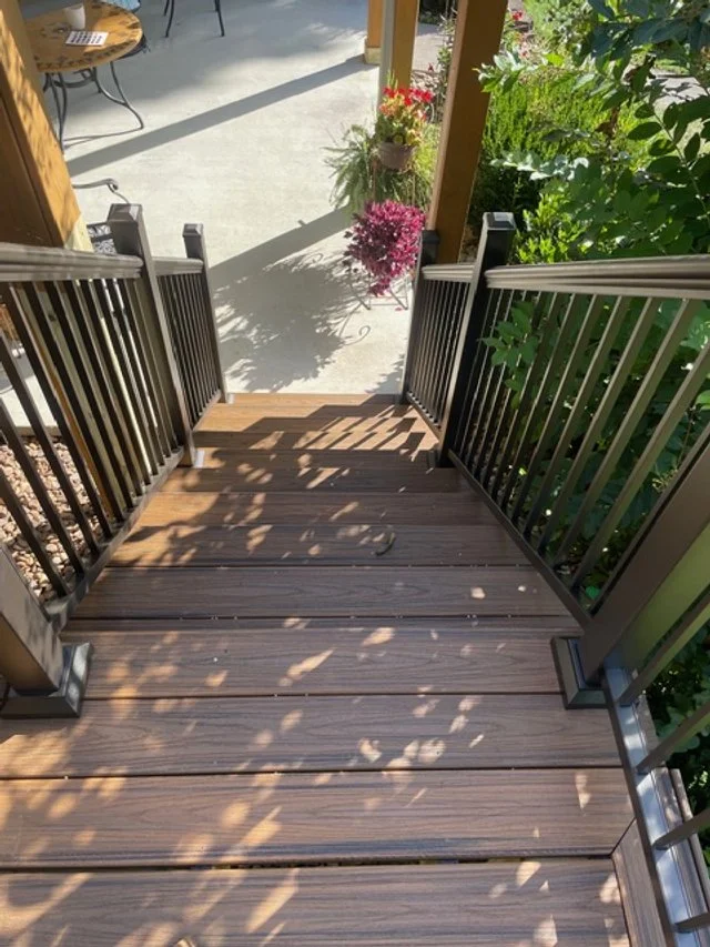 View of a wooden outdoor staircase with black metal railings leading from a patio to a garden area.