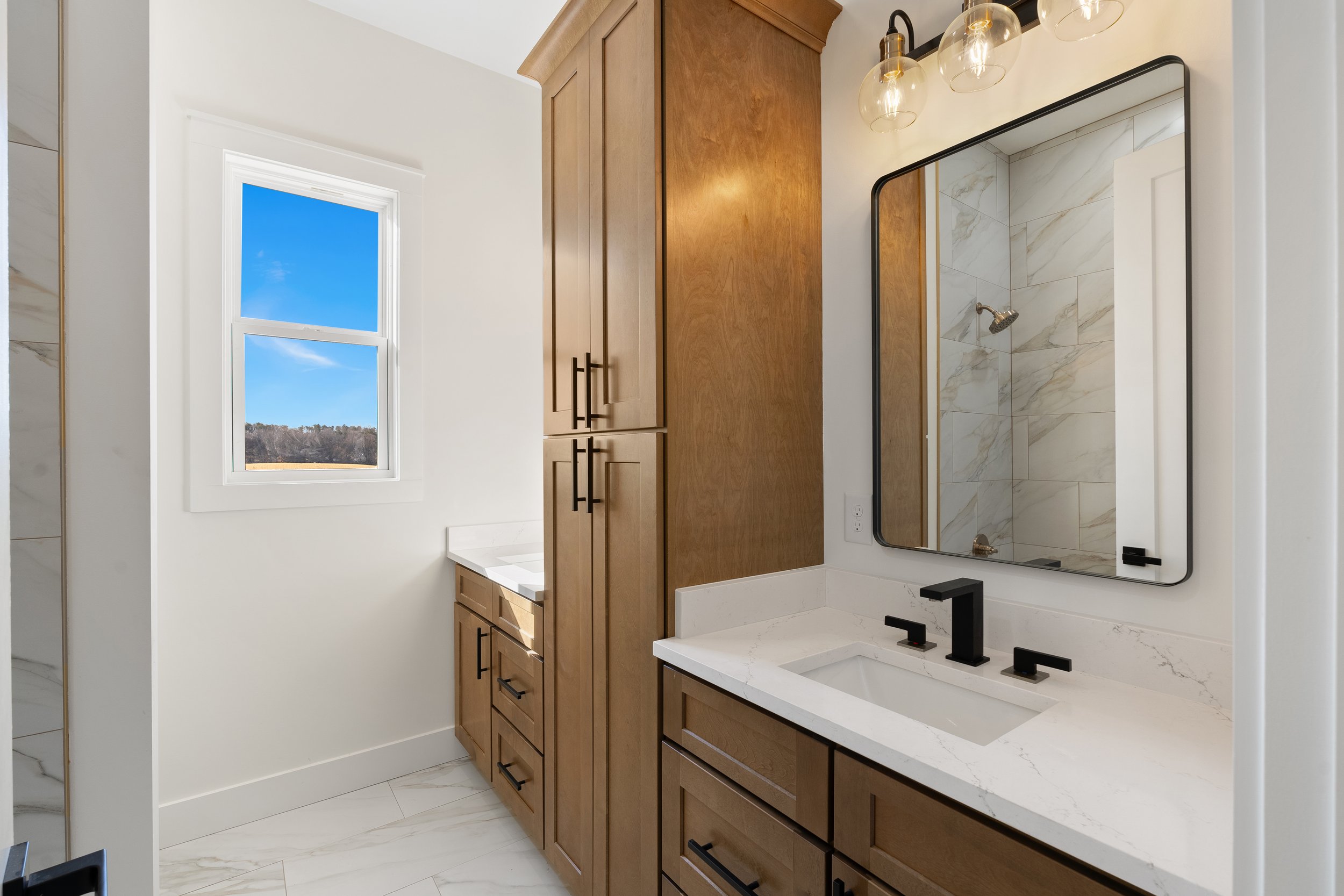 Modern bathroom with white walls, a wooden cabinet, a large mirror, a white marble countertop with a black faucet, a window showing a blue sky with clouds and landscape, and a shower area with marble tiles and a showerhead.