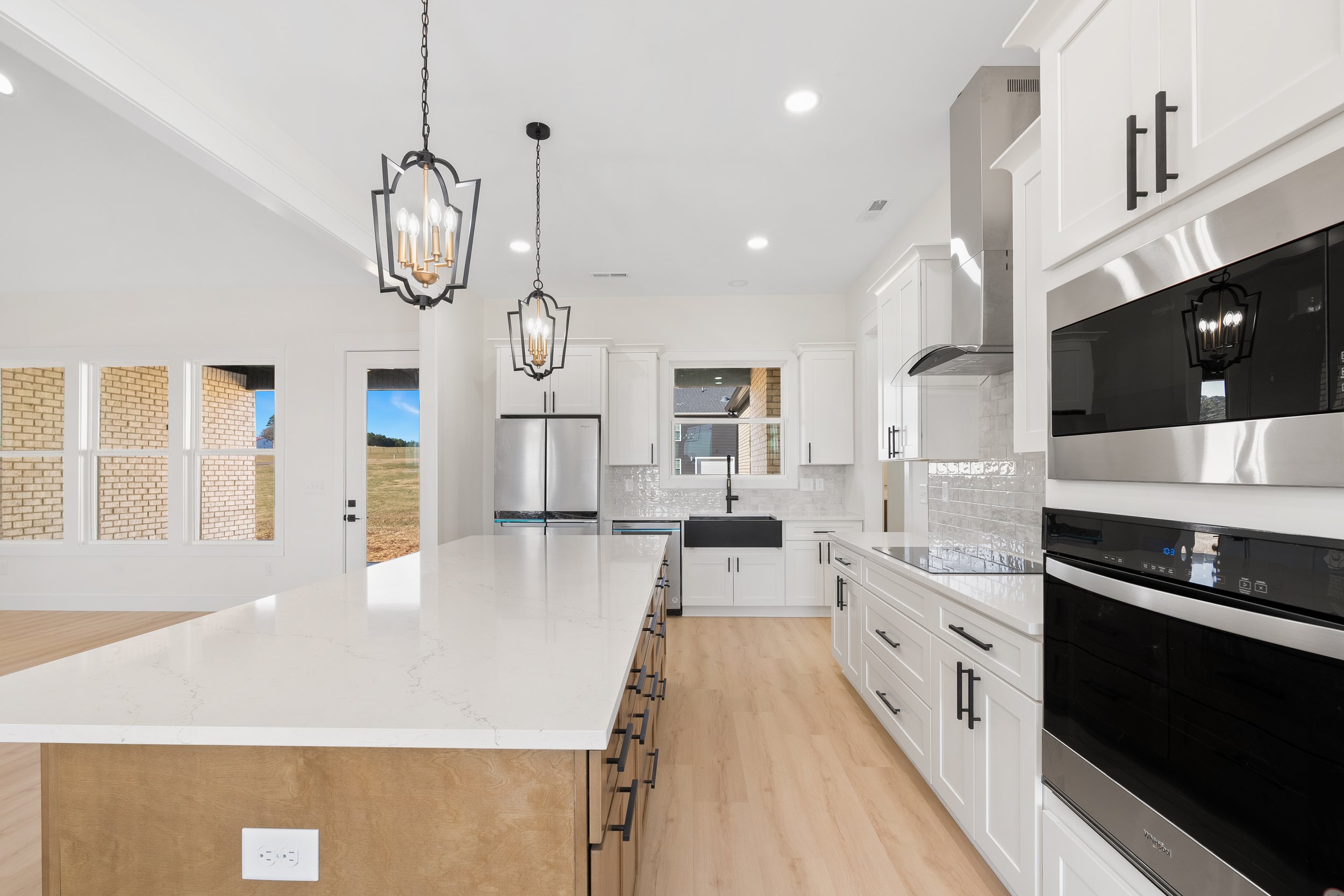 Modern kitchen with white cabinets, black hardware, stainless steel appliances, a black farmhouse sink, and pendant lights over a large white island.