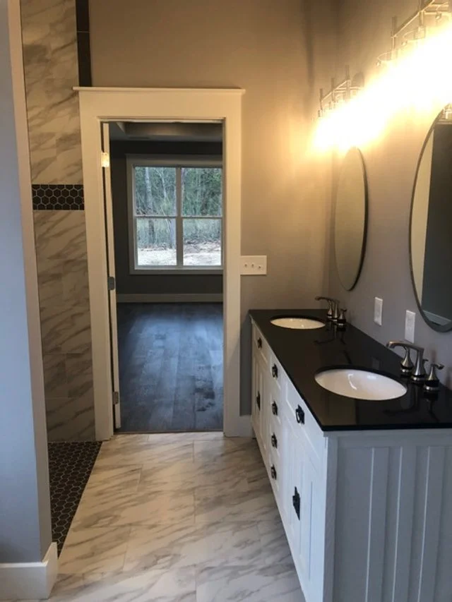 Bathroom with dual sinks, black countertops, oval mirrors, and a view into a room with large window and woods outside.