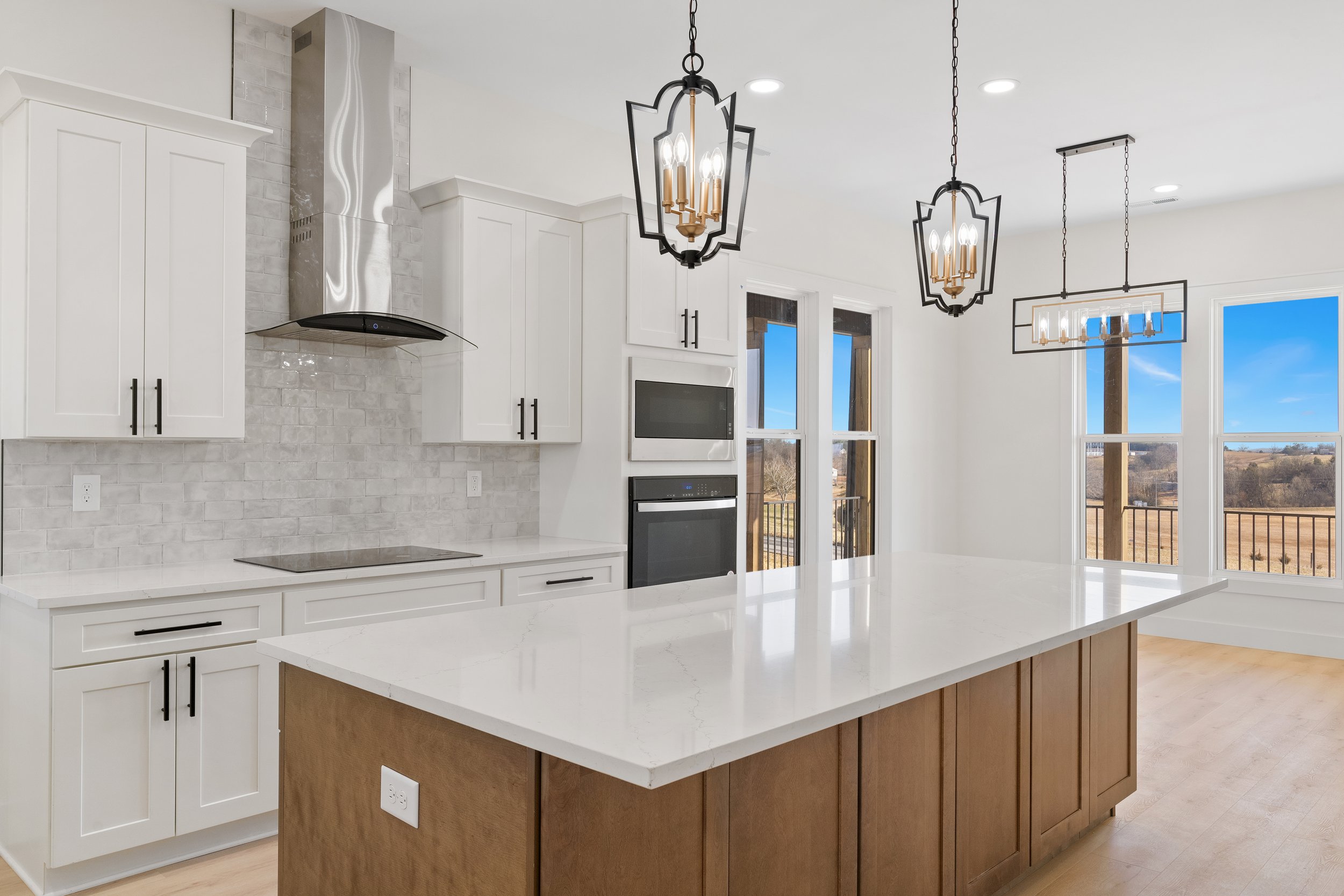 Modern kitchen with white cabinets, a marble island, black hardware, a stainless steel range hood, and large windows showing a scenic outdoor view.