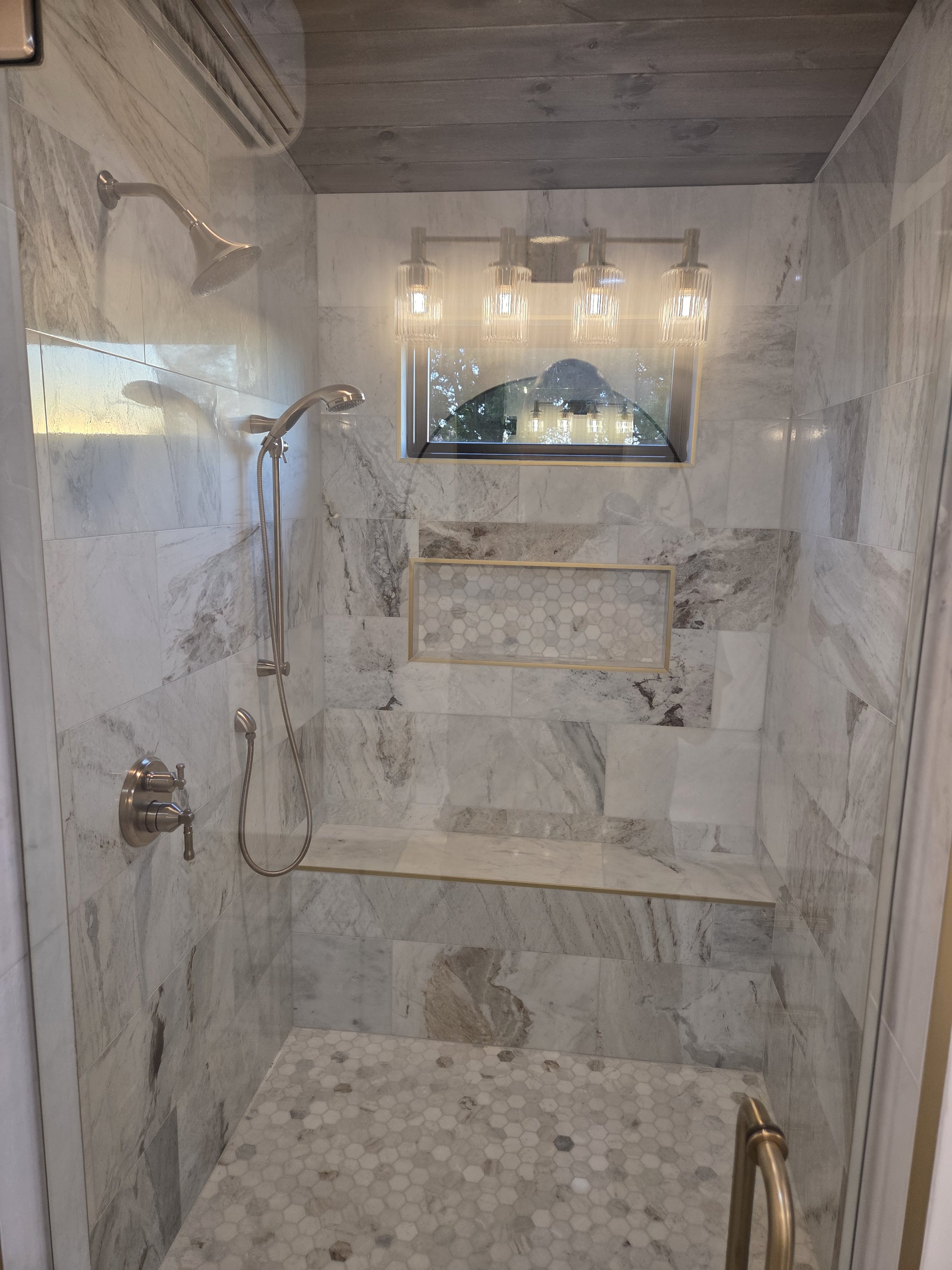 A modern shower with marble tiles on the walls and floor, two shower heads, a window with reflections, a ceiling light fixture, and a built-in bench.