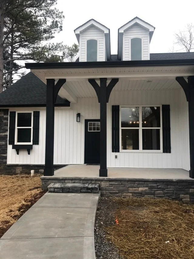 Front porch of a new white house with black trim, two small dormer windows on the roof, and a concrete walkway leading to the front door.