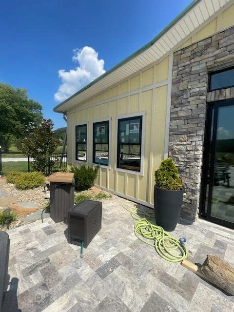 Exterior of a yellow house with stone accents, three windows, and a glass door. Outdoor patio area with potted plants, a small table, a waste bin, and garden hose on a cloudy day.