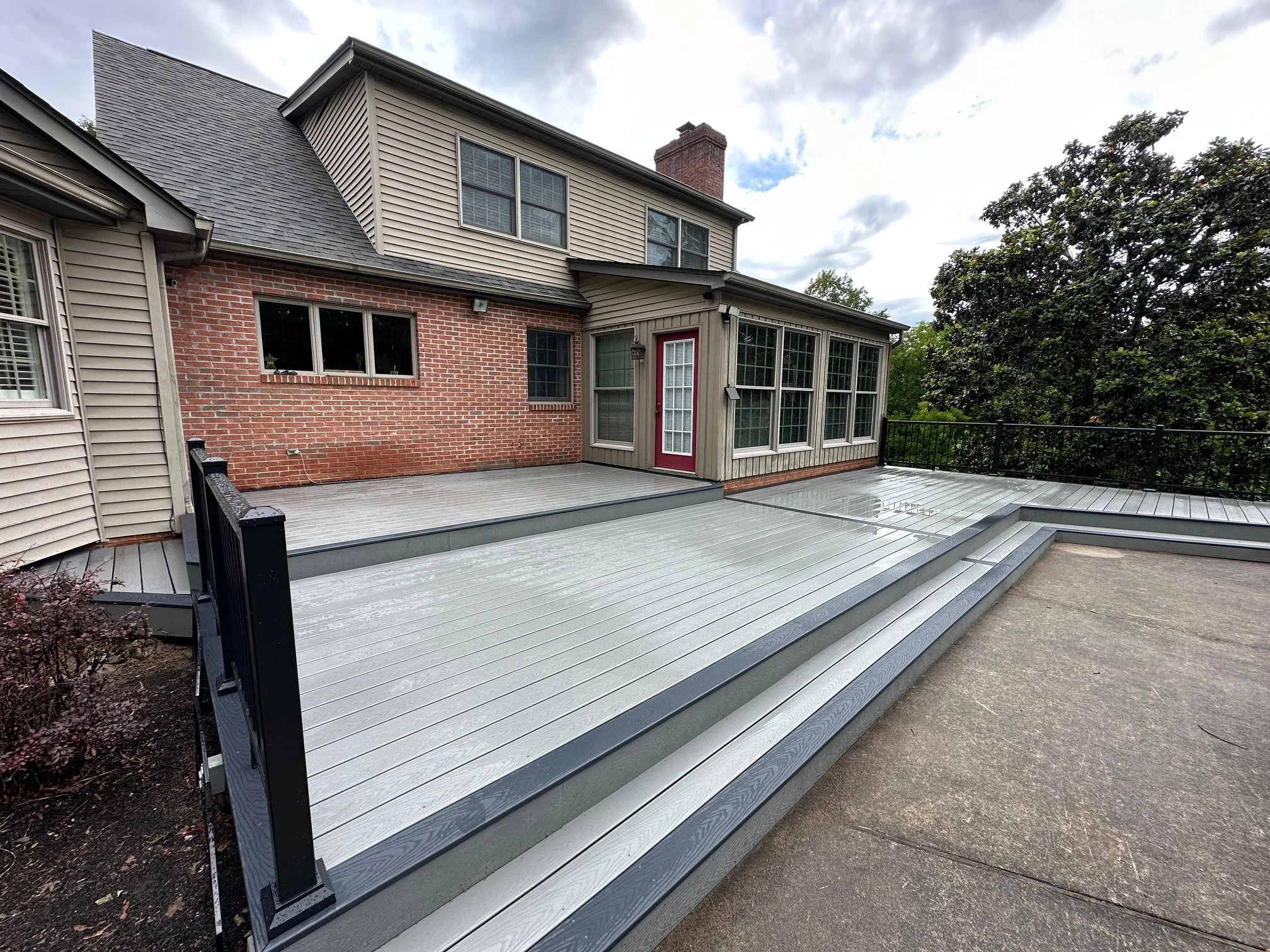 Backyard deck with gray metal flooring, black railings, and a house with brick and beige siding. The house has several windows and a red door.