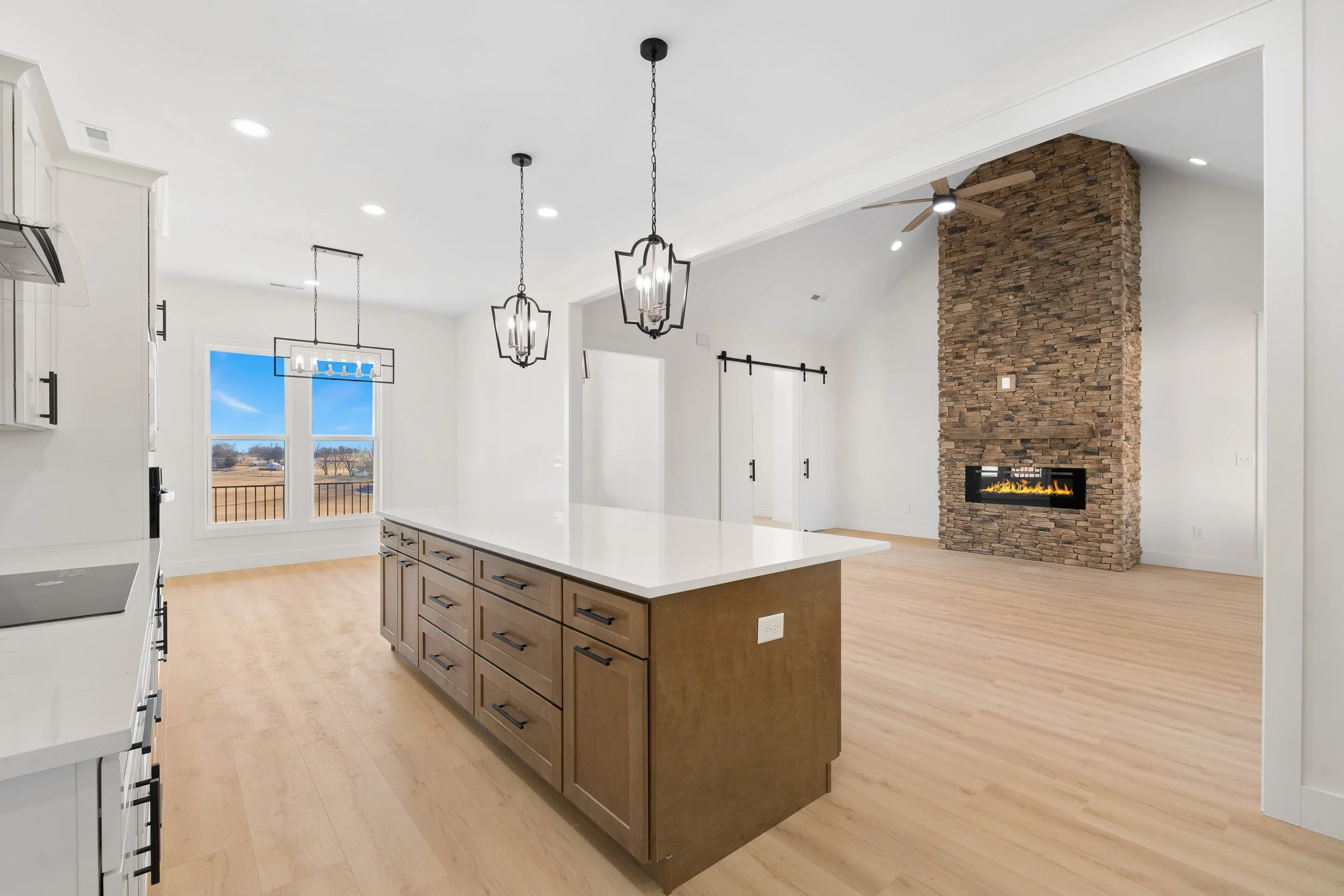 Modern open-concept kitchen and living room with a stone fireplace, large window, light hardwood floors, and contemporary lighting fixtures.