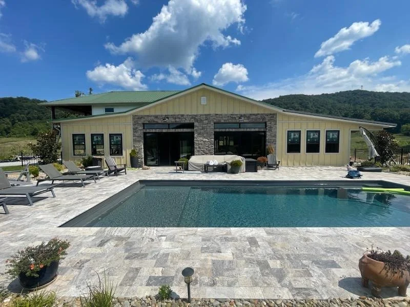 Modern backyard with a swimming pool, lounge chairs, patio furniture, and a house with large windows and a stone facade, set against a background of green hills and blue sky with clouds.