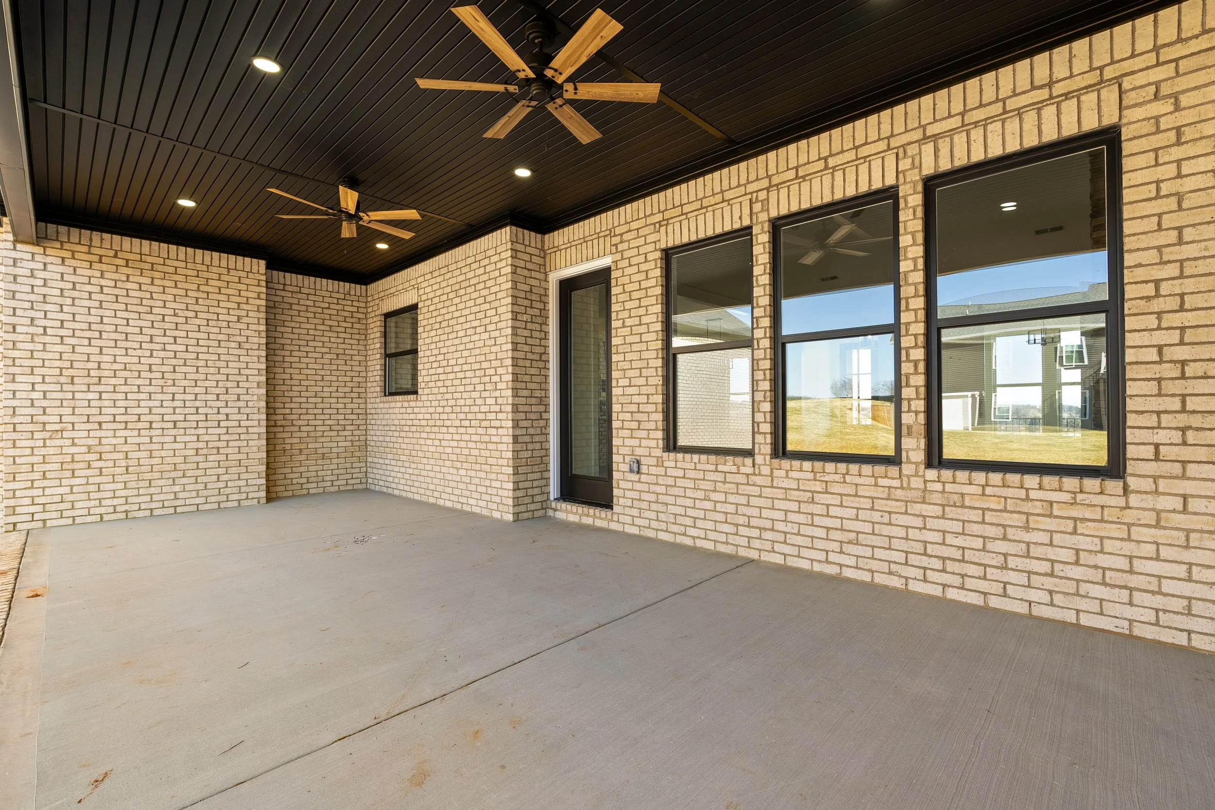 Covered patio with beige brick walls, black-framed windows, concrete flooring, black ceiling with ceiling fans and recessed lighting.