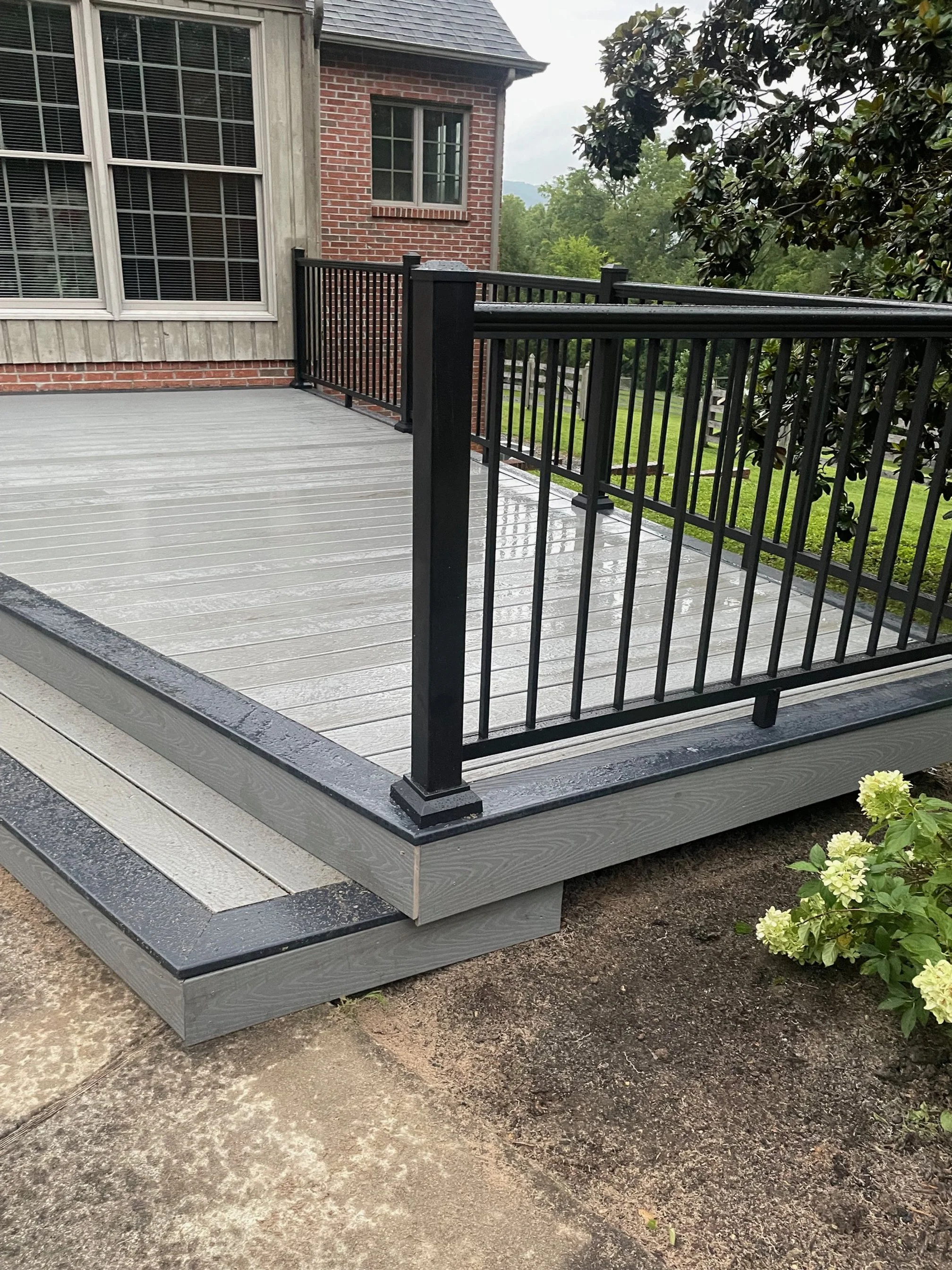A newly built outdoor deck with gray wooden flooring and black metal railing, attached to a house with a brick and wood exterior, surrounded by greenery and flowering bushes.