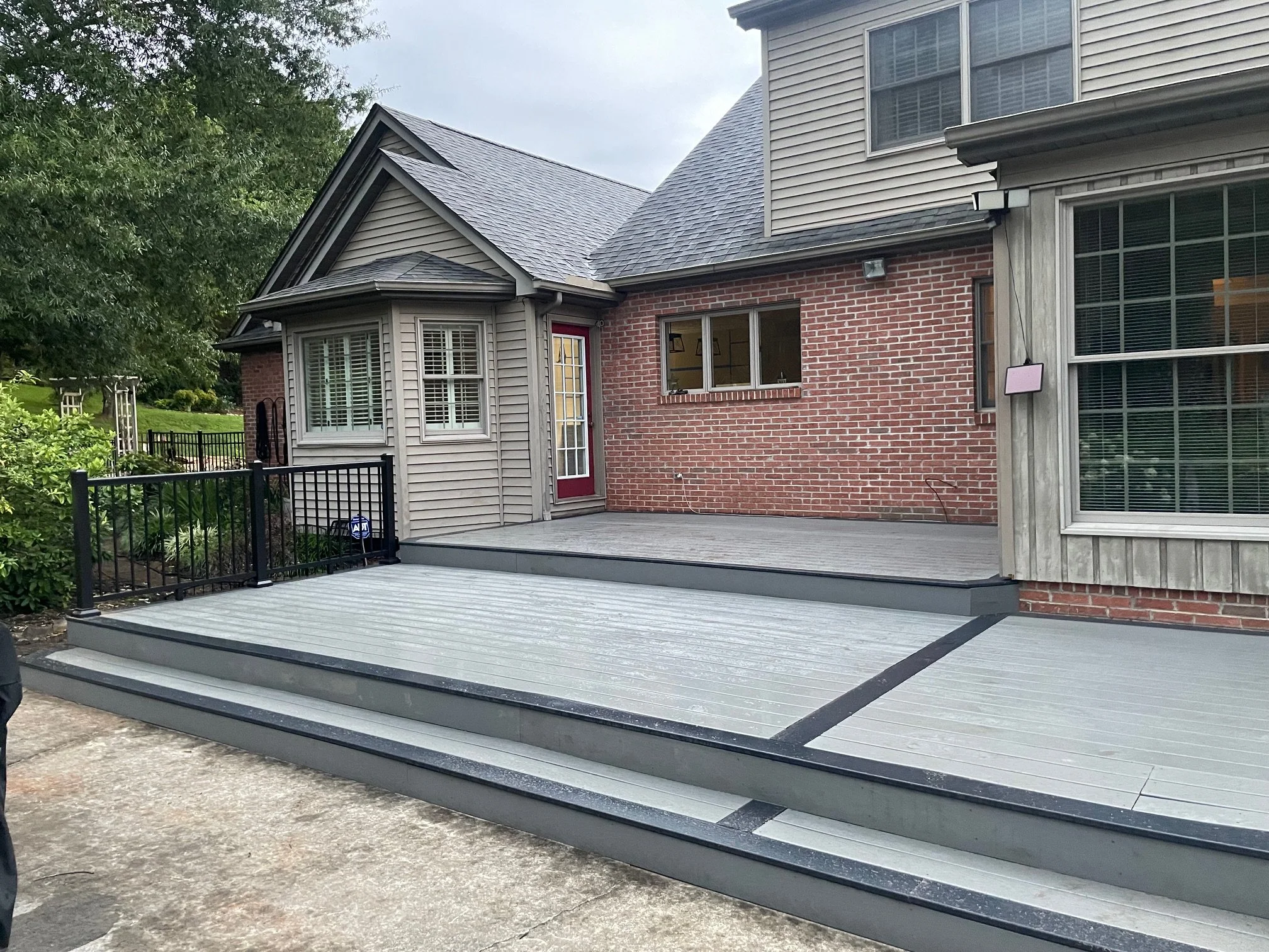 Newly installed multi-level composite deck with black metal railing attached to the back of a brick and siding house, surrounded by greenery.