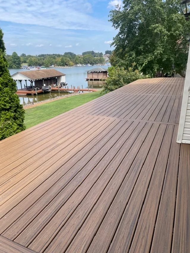 View of a wooden deck overlooking a lake with docked boats and boathouses, grassy yard, trees, and a partly cloudy sky.