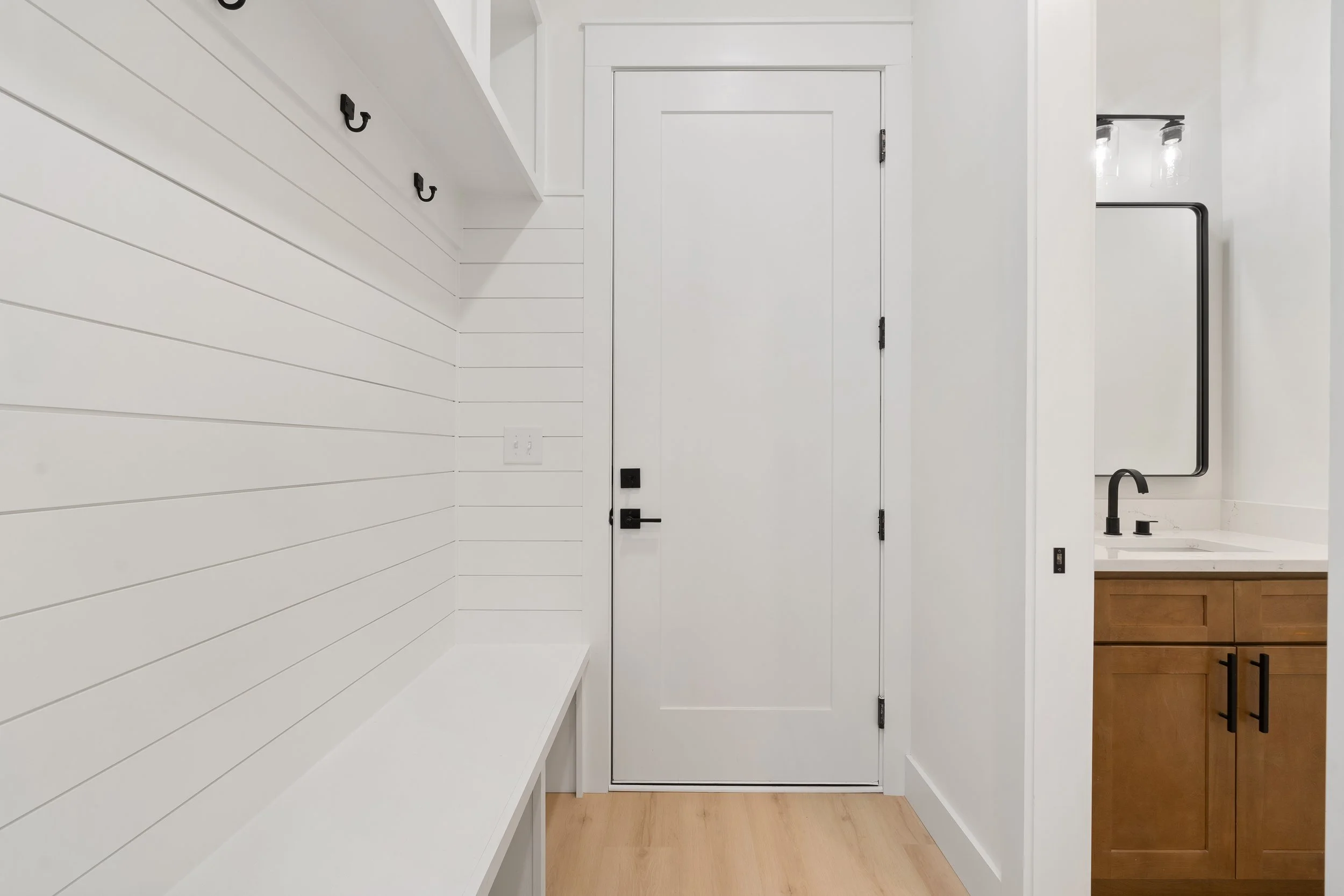 A modern entryway with white shiplap walls, black hooks, a white door, and a small bathroom with a mirror and wooden cabinet.