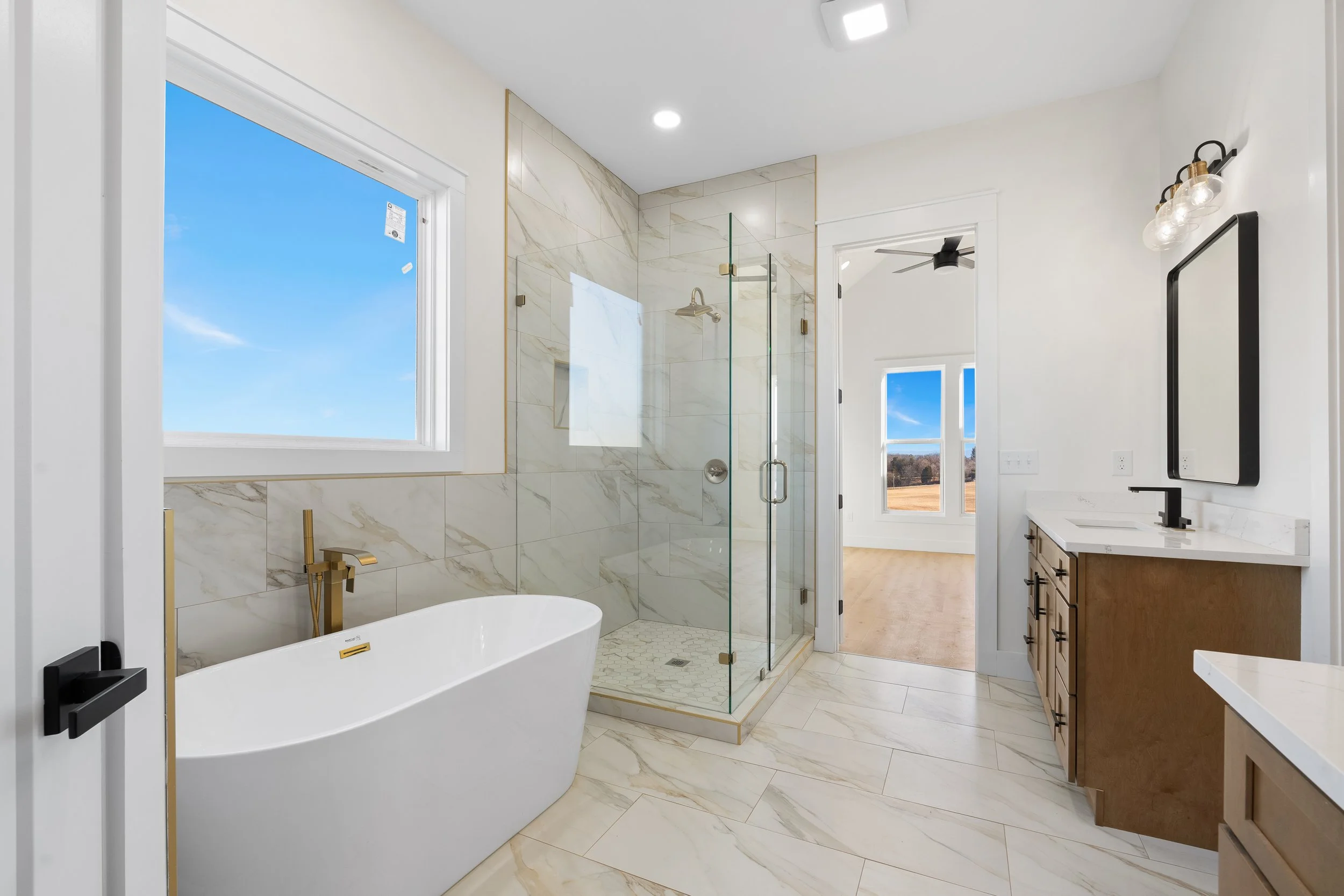 Modern bathroom with a bathtub under a window, a glass-enclosed shower, a wooden vanity with a white countertop, and large windows showing an outdoor landscape.
