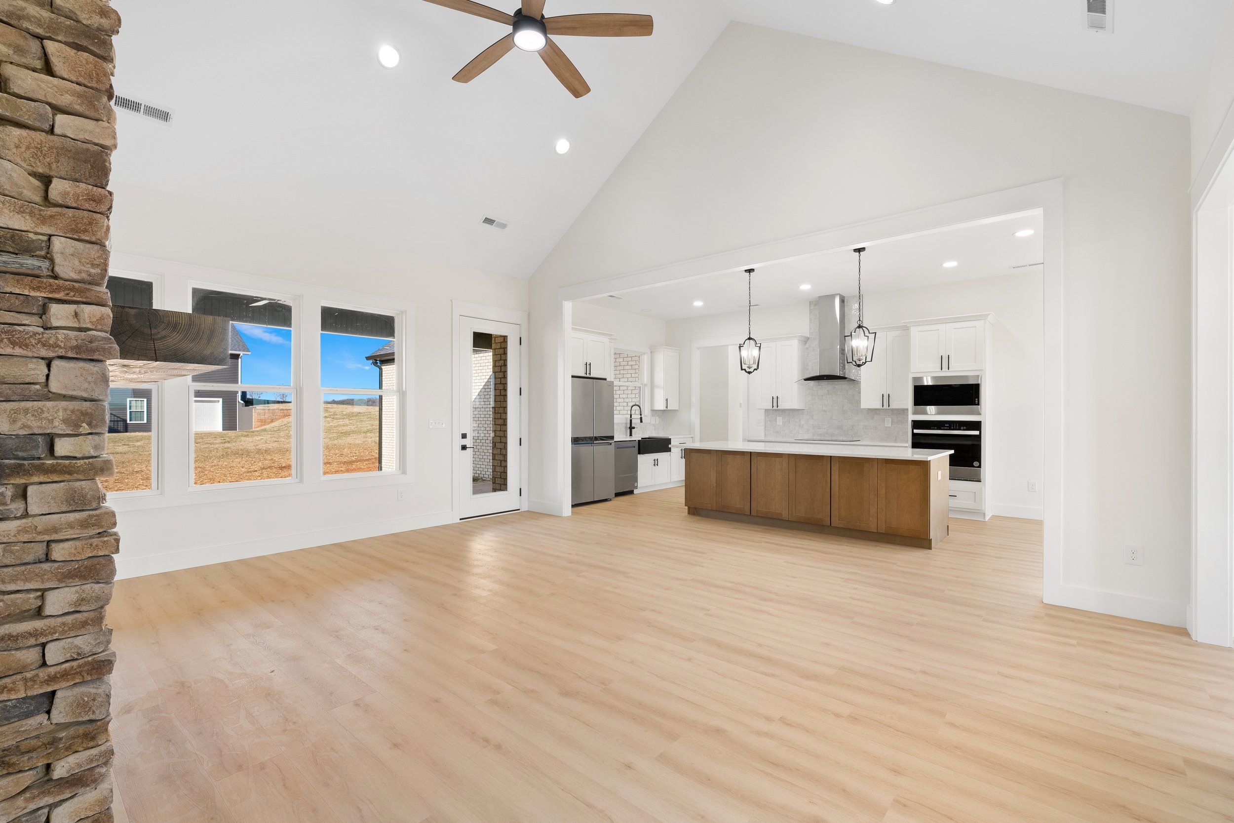 Empty modern kitchen and living area with large windows, wooden floor, kitchen island, white cabinets, and stainless steel appliances.