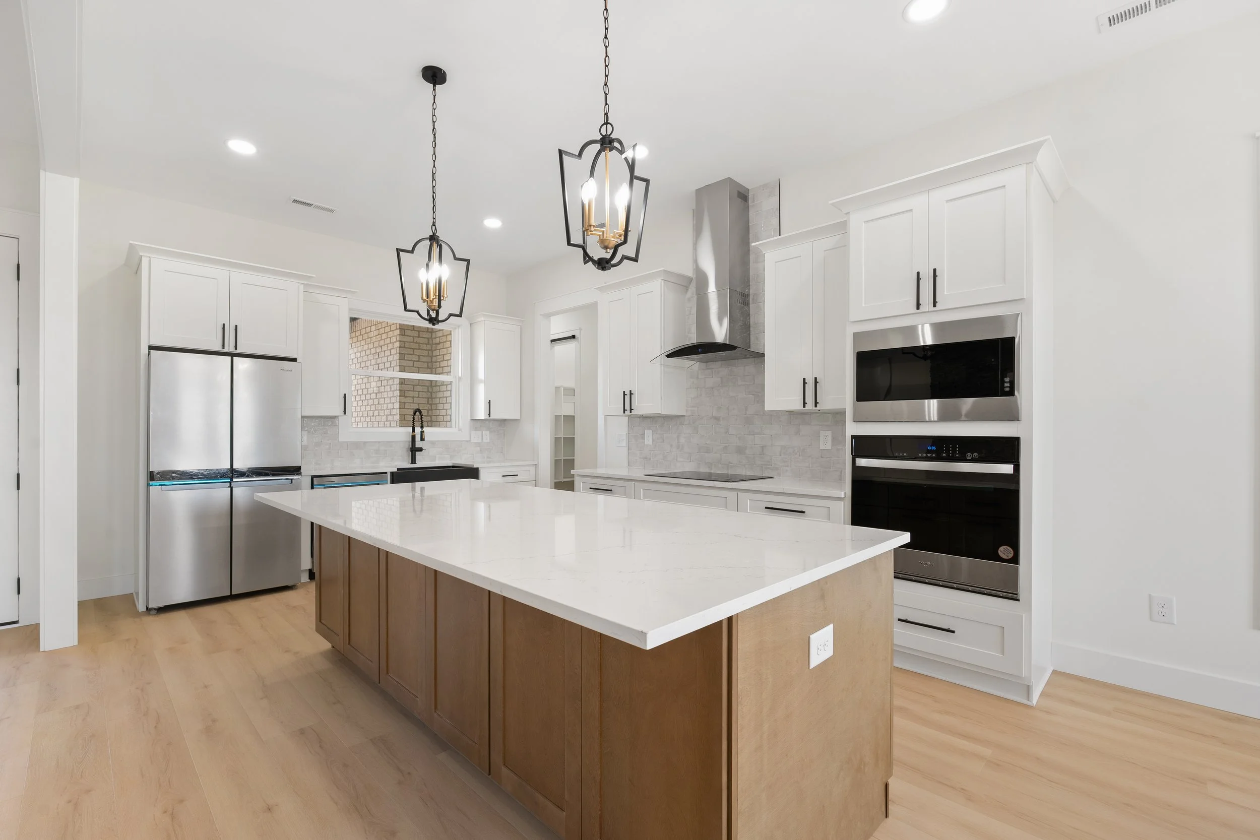 Modern white kitchen with an island, stainless steel refrigerator, oven, microwave, white cabinets, black handles, brick backsplash, black sink, black faucet, pendant lights, hardwood floors, and a window above the sink.