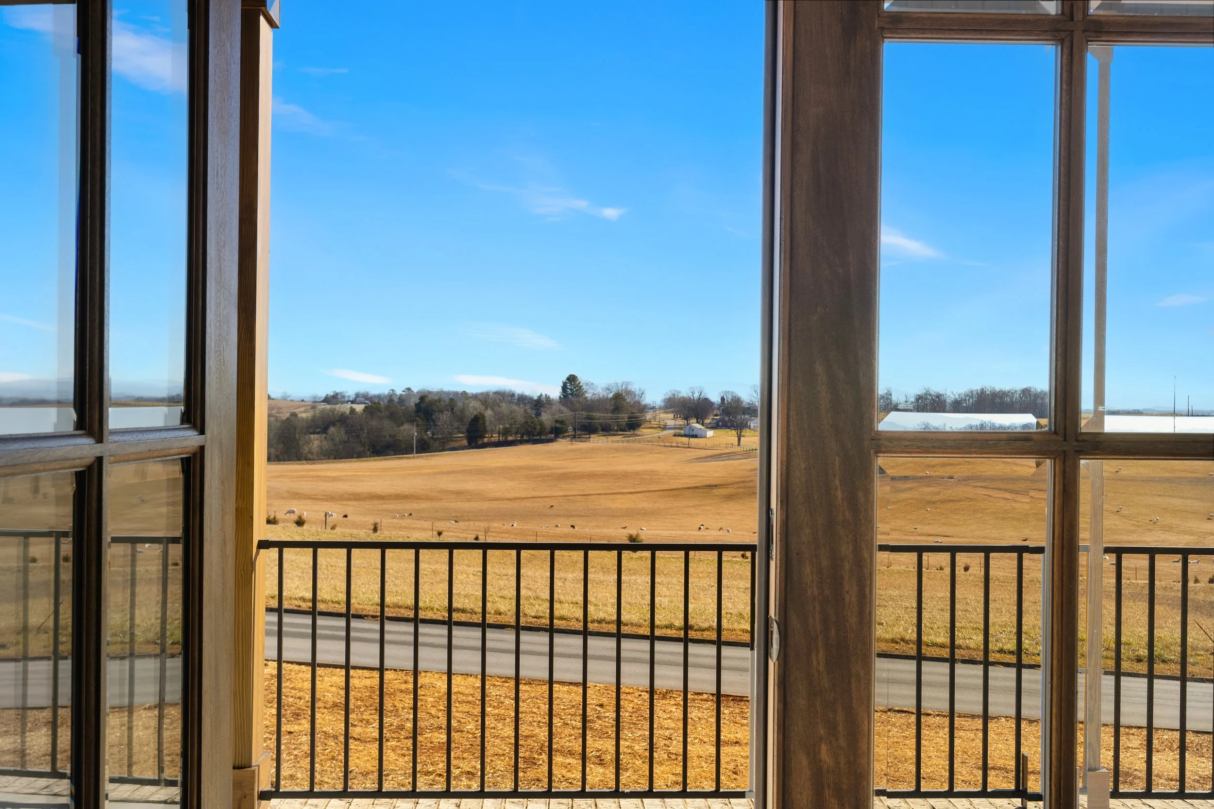 View of a rural landscape through an open window with wooden frames and black railing, showing a field, trees, and a blue sky.