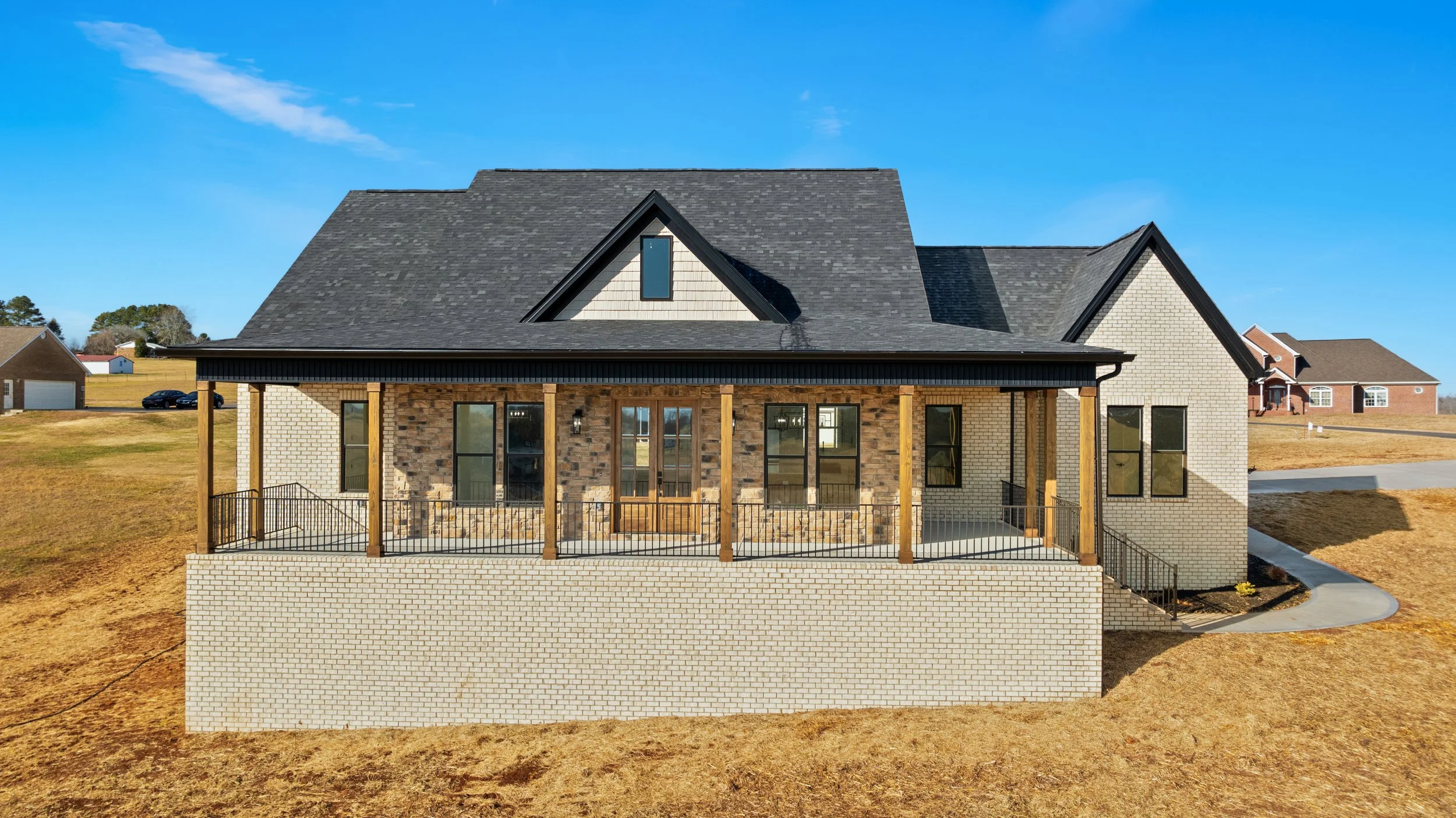 Newly constructed house with a front porch, brick and stone exterior, multiple large windows, and a sloped black roof, situated in a suburban neighborhood with other houses visible in the background under a clear blue sky.