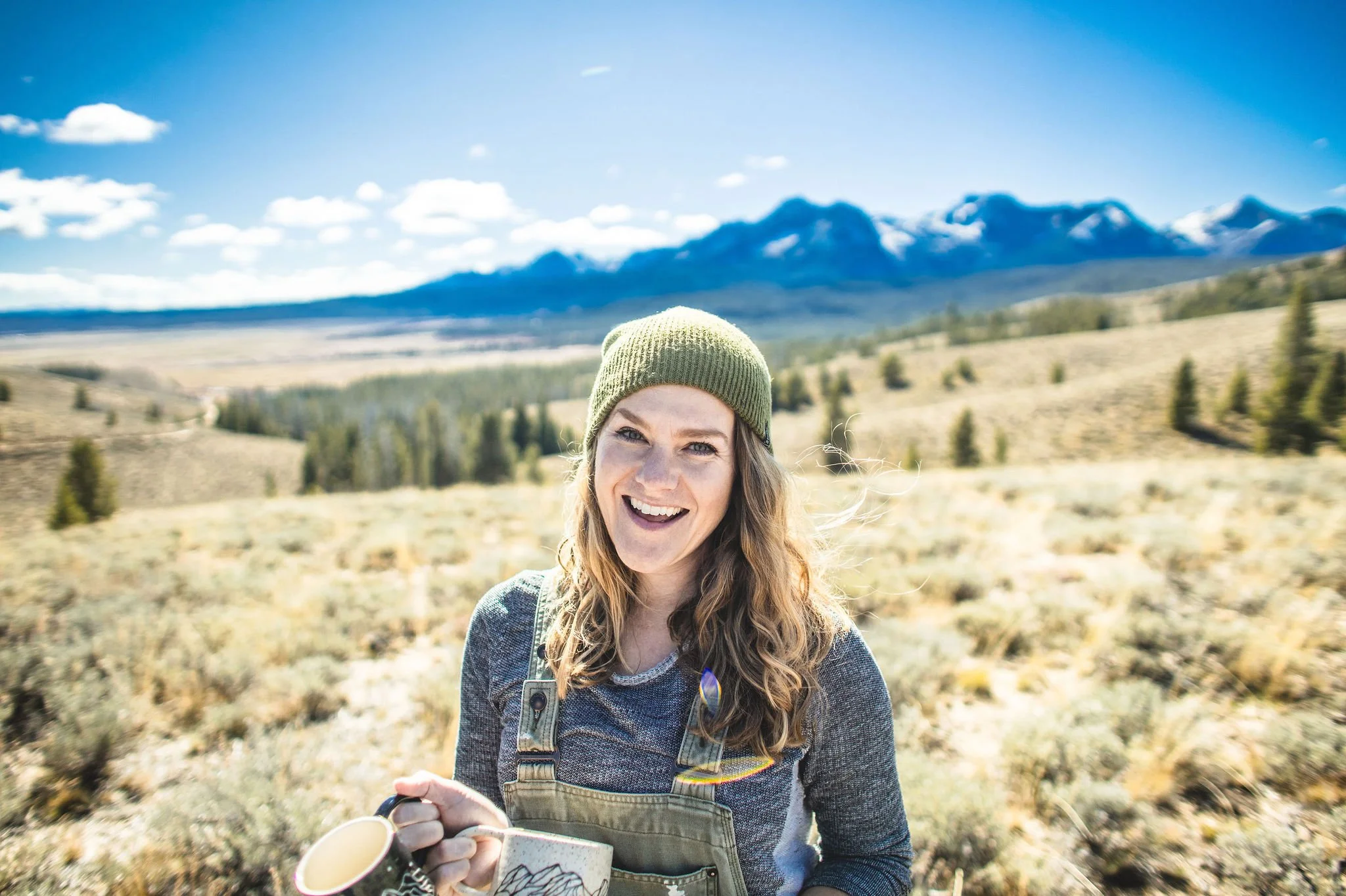 A woman with curly hair wearing a green beanie and overalls, smiling and holding a mug in a mountainous landscape with clear blue skies, sparse trees, and yellowish grass.
