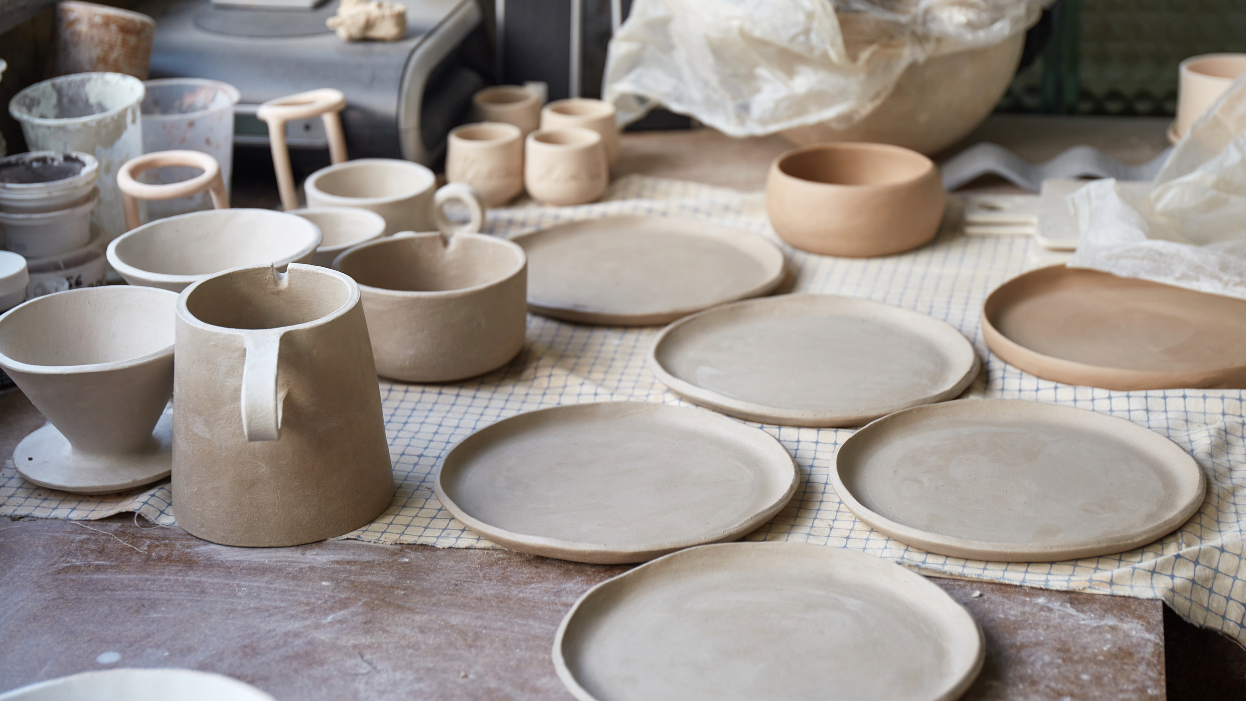 Unfinished pottery pieces including bowls, plates, and a teapot, sitting on a cloth-covered worktable in a ceramic studio.