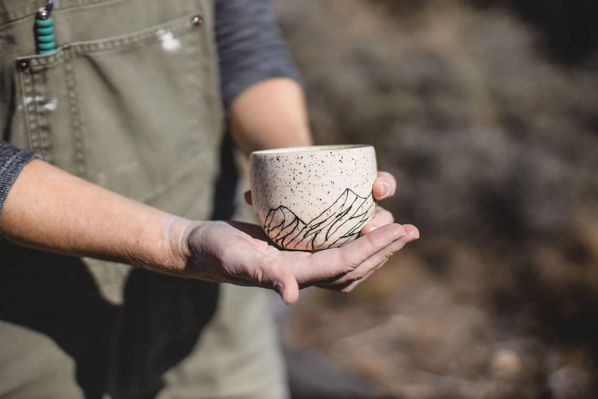 Person holding a ceramic cup with a mountain design, outdoors.