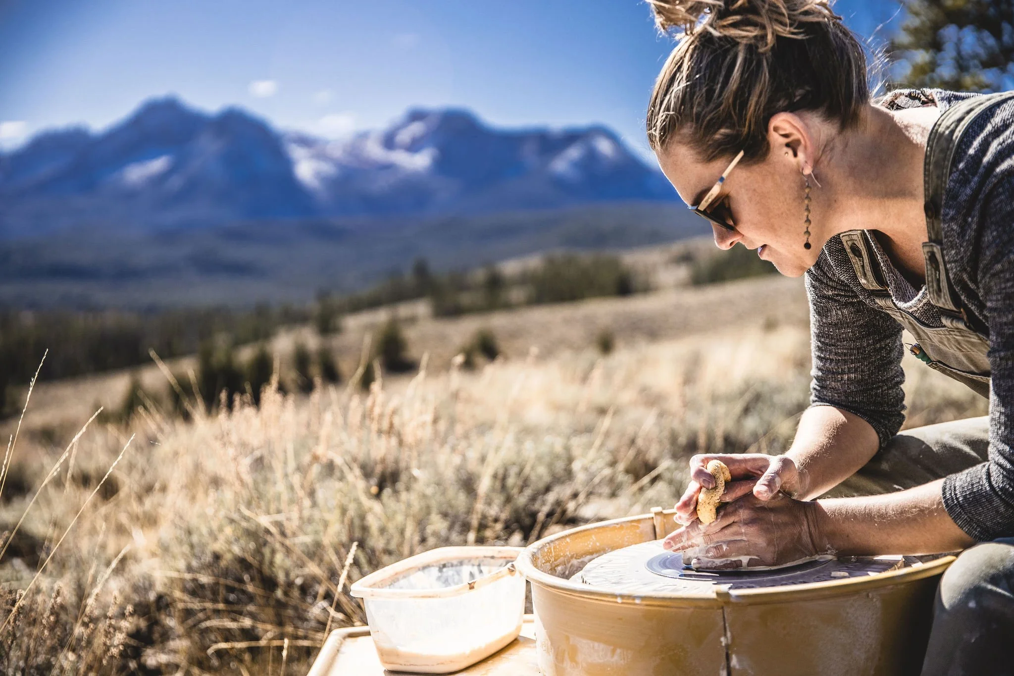 A woman working on a pottery wheel outdoors in a field with mountains in the background.