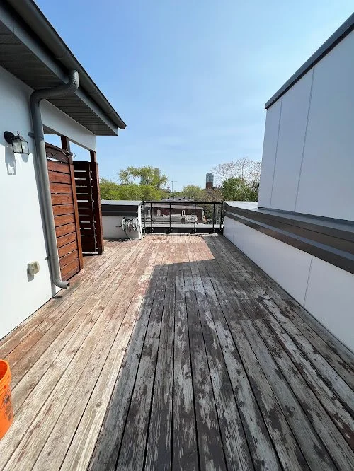 Rooftop balcony with wooden floor, partial wall, some outdoor furniture, and trees in the distance under a clear blue sky.