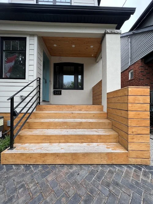 Pressure treated deck refinishing in Toronto delivering smooth, uniform stain finish. Wooden front porch stairs leading to a house entrance, with a black metal railing on one side, a white exterior wall, and a wooden ceiling over the porch.
