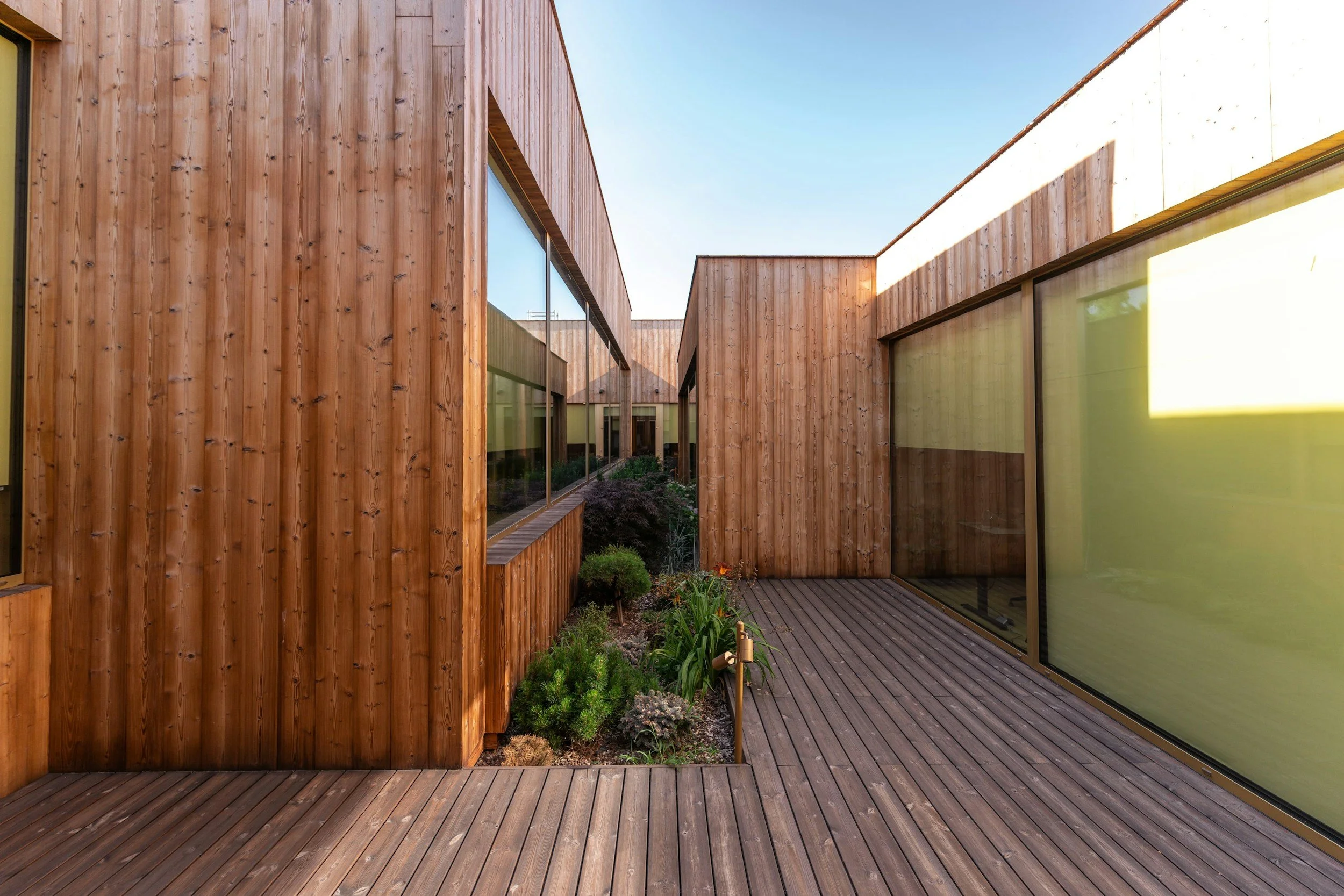 Outdoor wooden deck area with wooden walls and large glass windows, neighboring a garden with small plants and shrubs, under a clear blue sky.