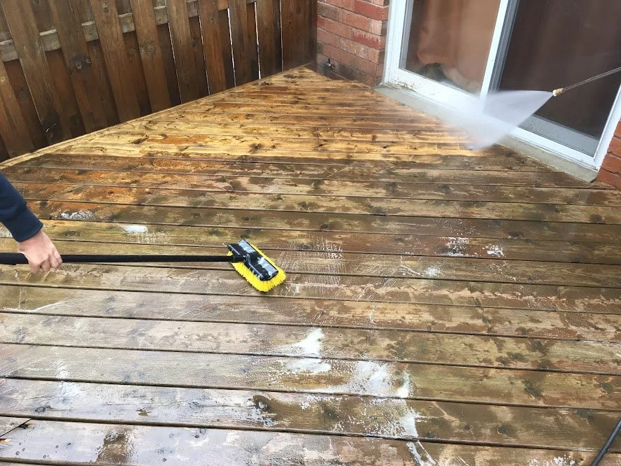A person cleaning a wooden deck with a power scrubber, water spray, and soap.