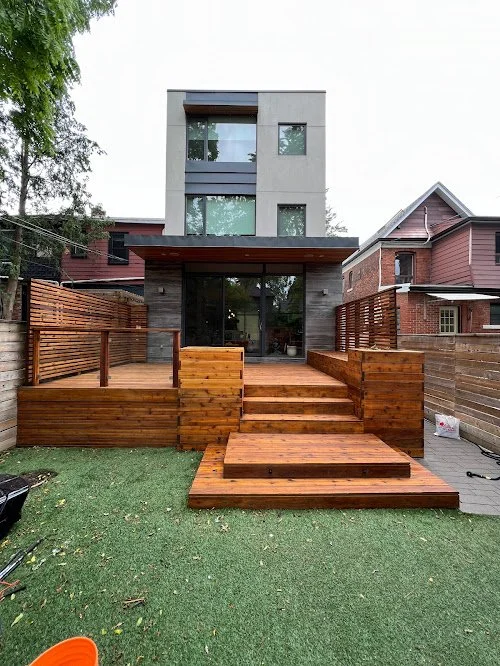 Cedar deck maintenance in the GTA extending lifespan with washing and protective sealing. Modern backyard with wooden deck, stairs, and fencing in front of a multi-story house with large glass doors.
