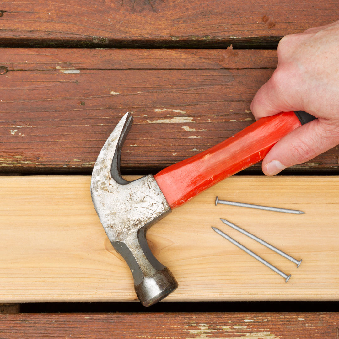 A person holding a hammer above a wooden board with two nails placed on it, ready for nailing.