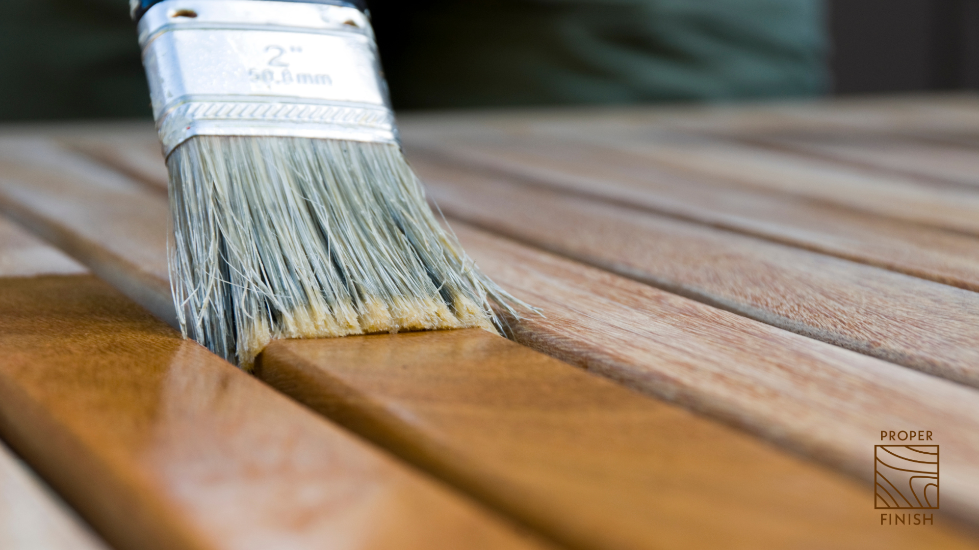 Close-up of a paintbrush applying a fresh coat of brown stain or varnish to a wooden surface, with the wood planks evenly coated.