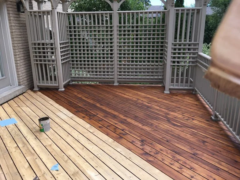 A deck under construction with partially stained wooden planks, a can of stain, and some blue painter's tape, enclosed by a grey wooden lattice fence.