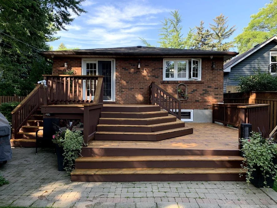 Newly renovated brick house with a wooden deck and stairs, surrounded by trees and neighboring houses, under a clear blue sky.