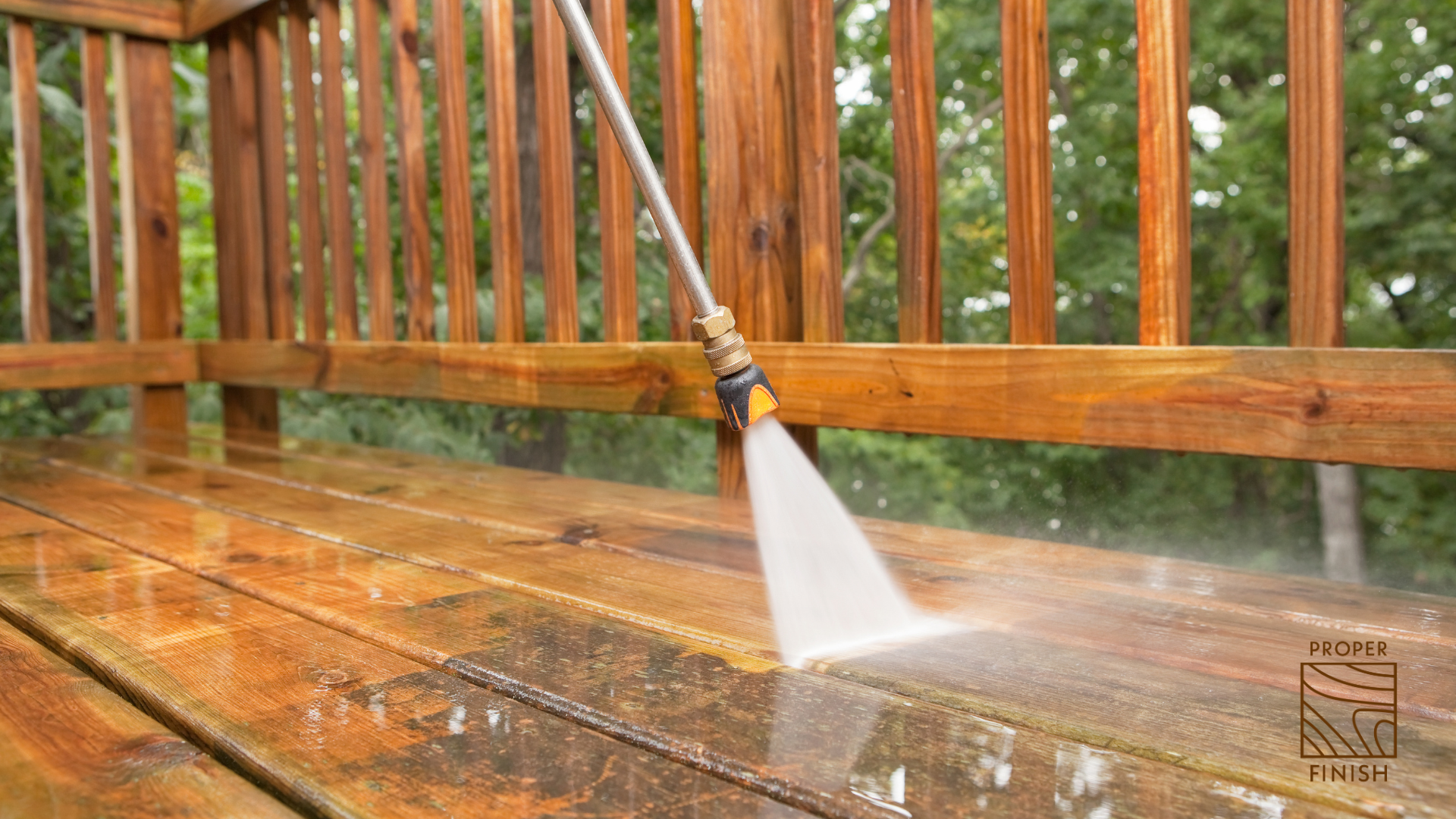 A garden hose with a spray nozzle watering a wooden deck outdoors, with trees in the background.