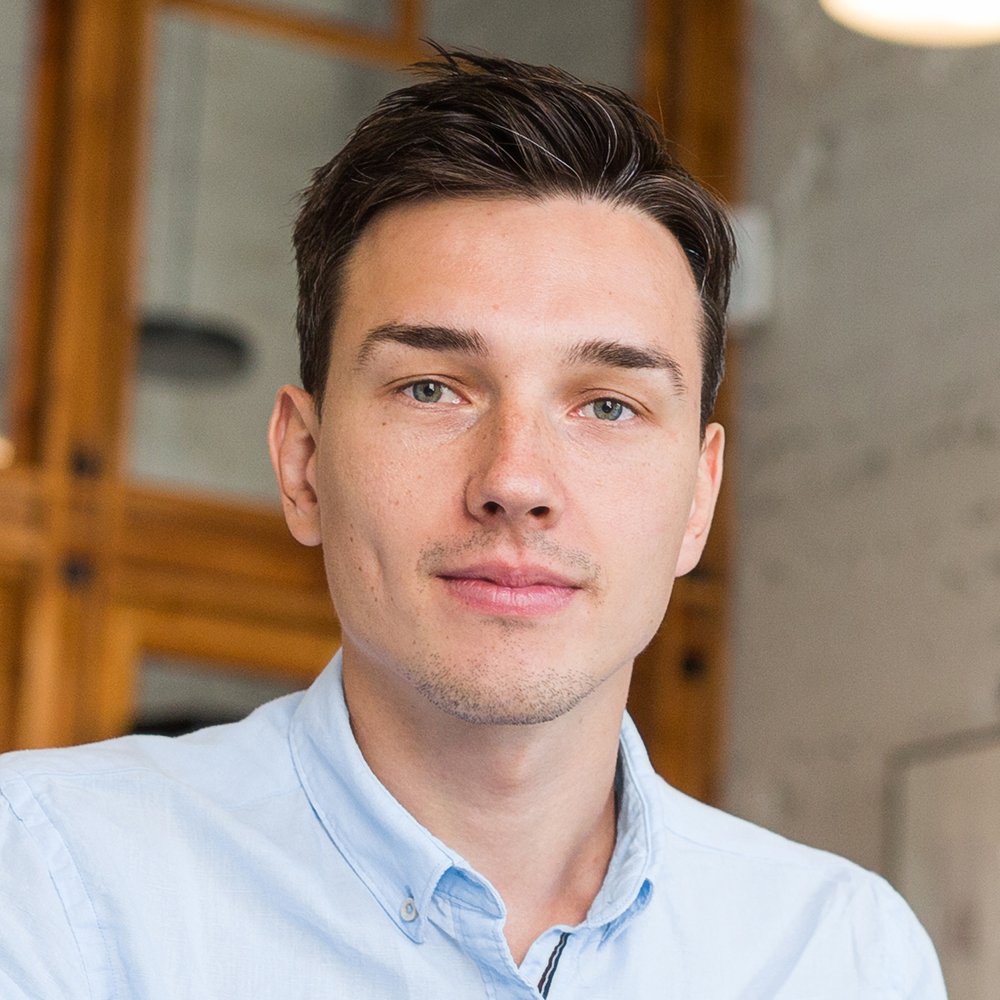 A young man with dark hair and blue eyes wearing a light blue collared shirt, looking at the camera with a neutral expression indoors.