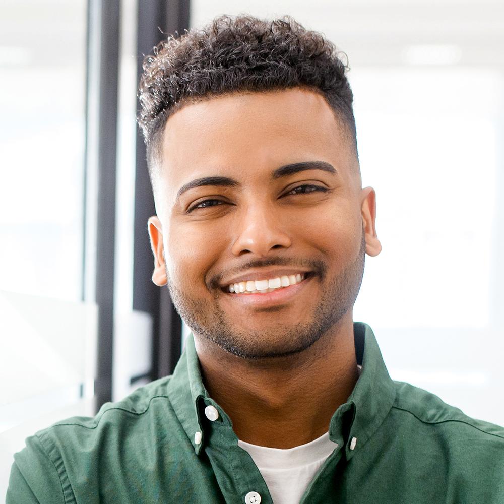 Close-up of a young man smiling, wearing a green button-up shirt with a white t-shirt underneath, indoors with natural light.