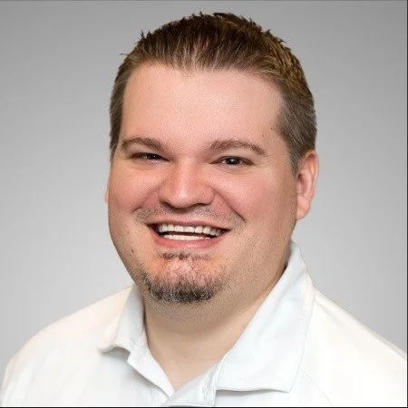 Portrait of a smiling man with short brown hair and a goatee, wearing a white shirt against a plain light gray background.
