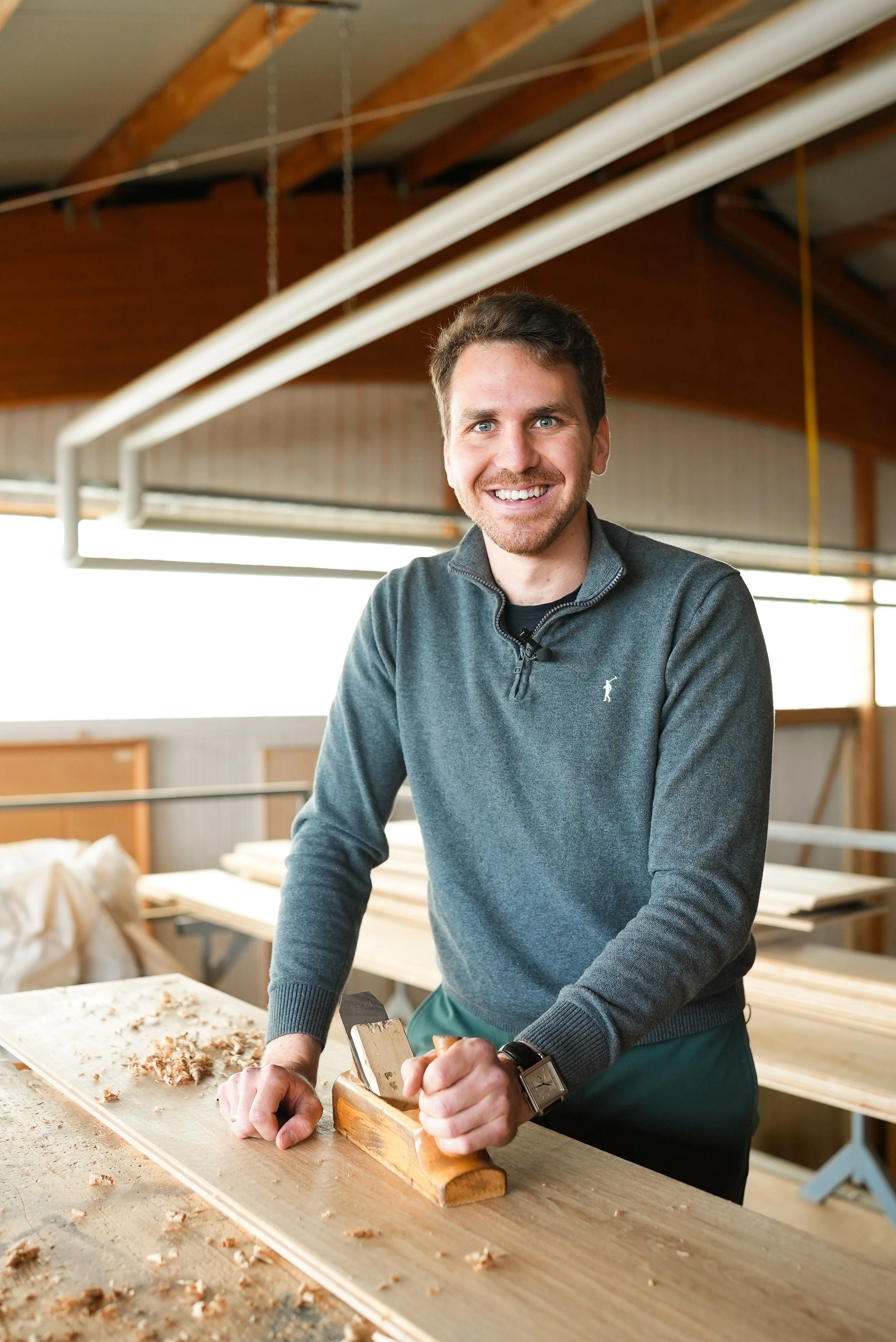 Ein Mann arbeitet in einer Tischlerei, er sägt Holz mit einer Handzugsäge und lächelt in die Kamera.