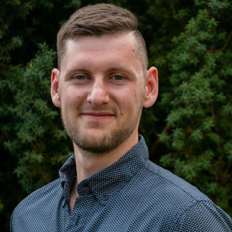 Junge Mann mit braunem Haar, kurzen Bart, dunklem Hemd, vor grünen Büschen.