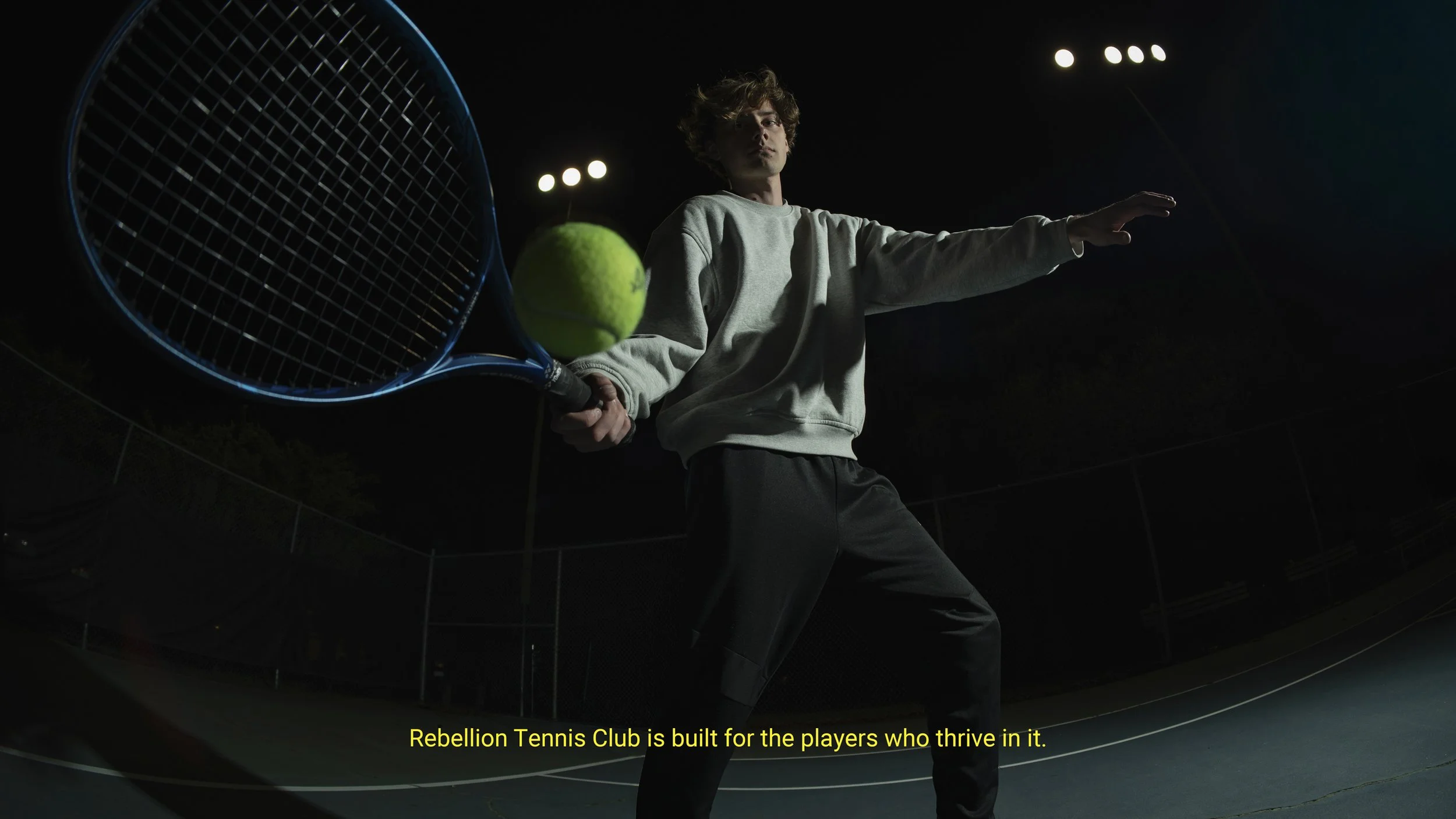 A person in a gray sweatshirt and black pants holding a tennis racket with a tennis ball near the racket, on a dark tennis court with bright lights in the background. Subtitles read: "Rebellion Tennis Club is built for the players who thrive in it."