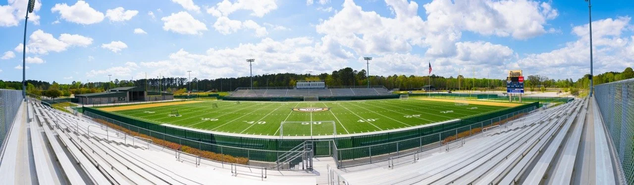 Photo of an empty football stadium with green field, bleachers, and a scoreboard under a partly cloudy sky.