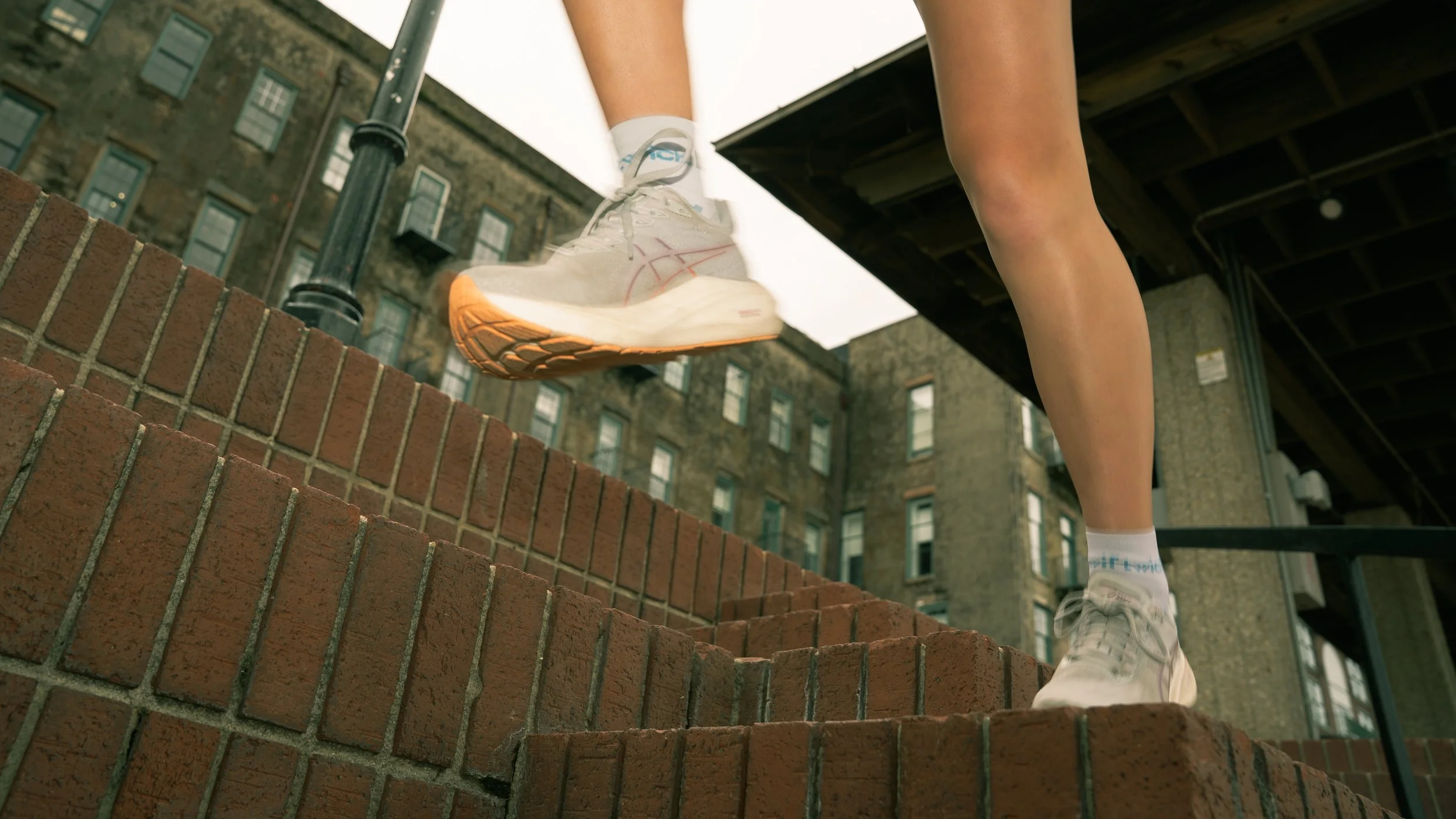 Low-angle view of a person running on a brick ledge, focusing on their white athletic shoes with orange accents, white socks, and black shorts, with an urban background of old brick buildings and a fire escape.
