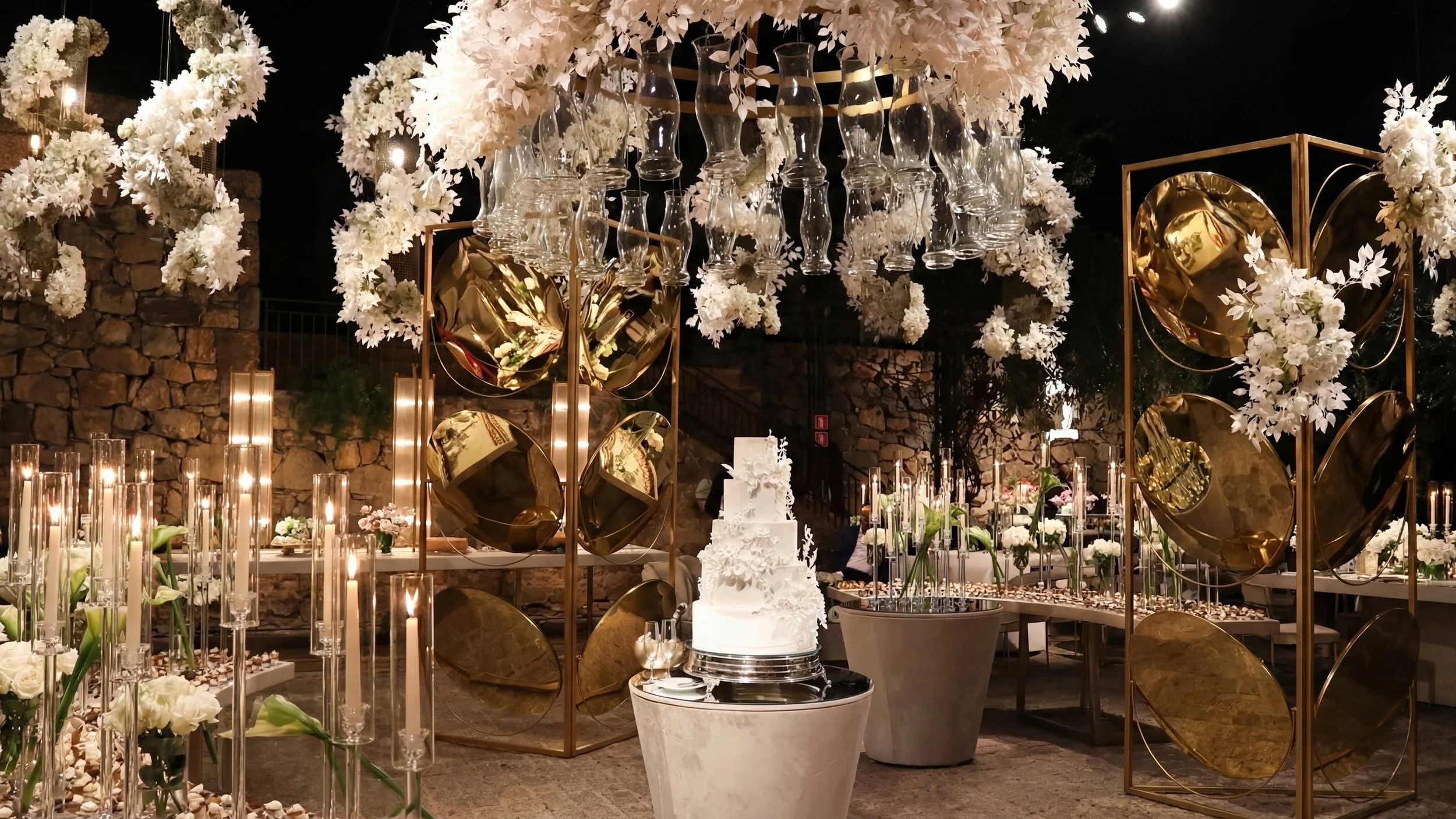 Decoração de festa com arranjos de flores brancas, velas em cilindros de vidro e um bolo branco decorado com flores, tudo com iluminação suave em ambiente interno com paredes de pedra.