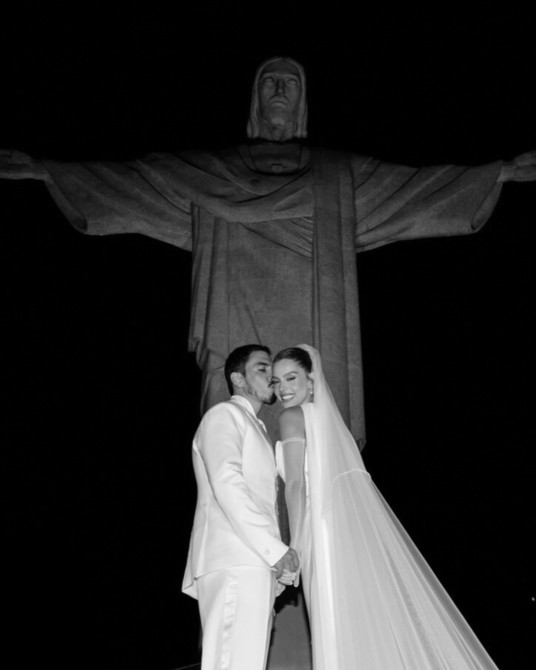 Casal de noivos na frente do Cristo Redentor durante a noite, sorrindo e segurando as mãos.