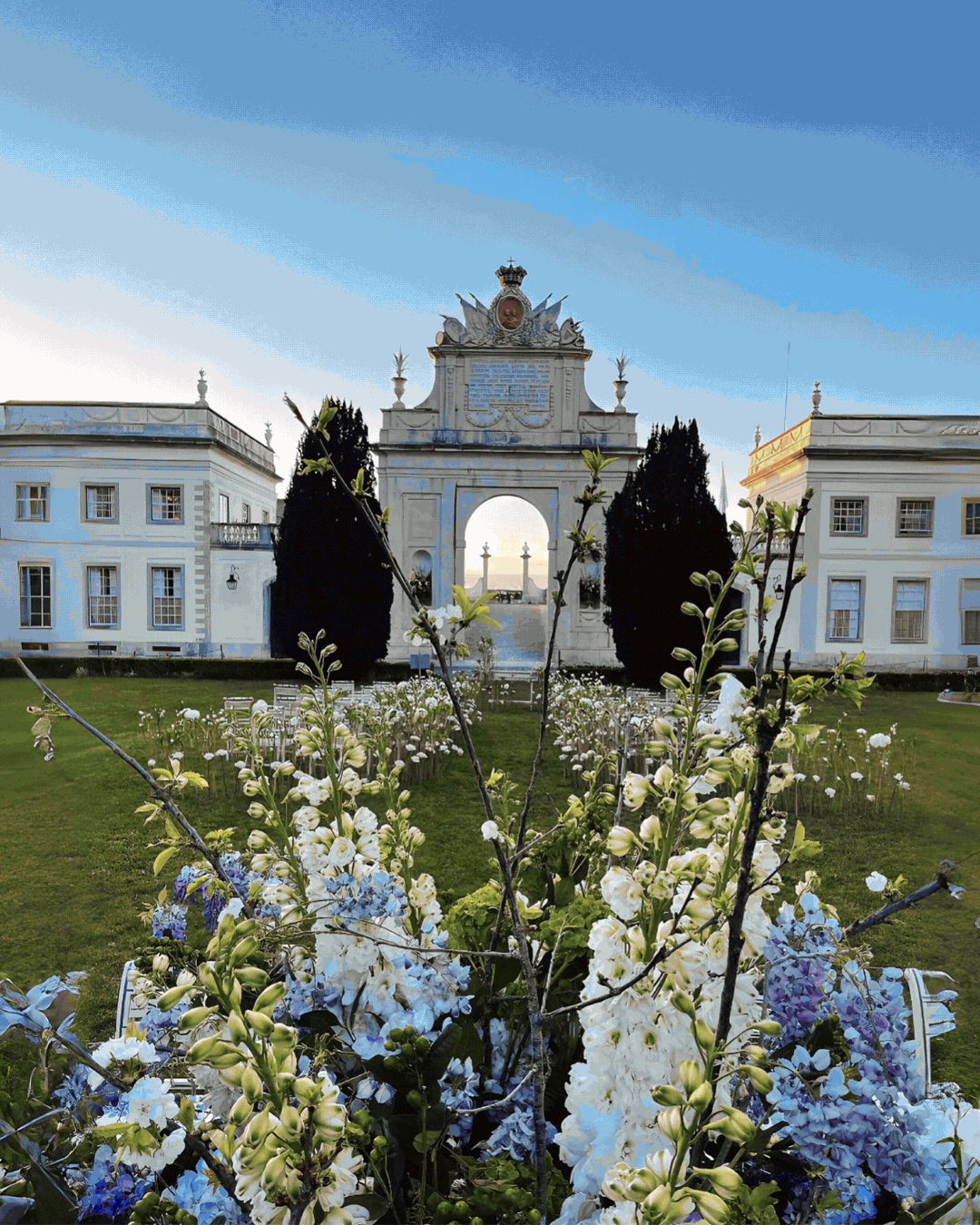 Arco do triunfo e edifícios históricos ao fundo, com flores brancas e lilás na frente, sob céu azul.