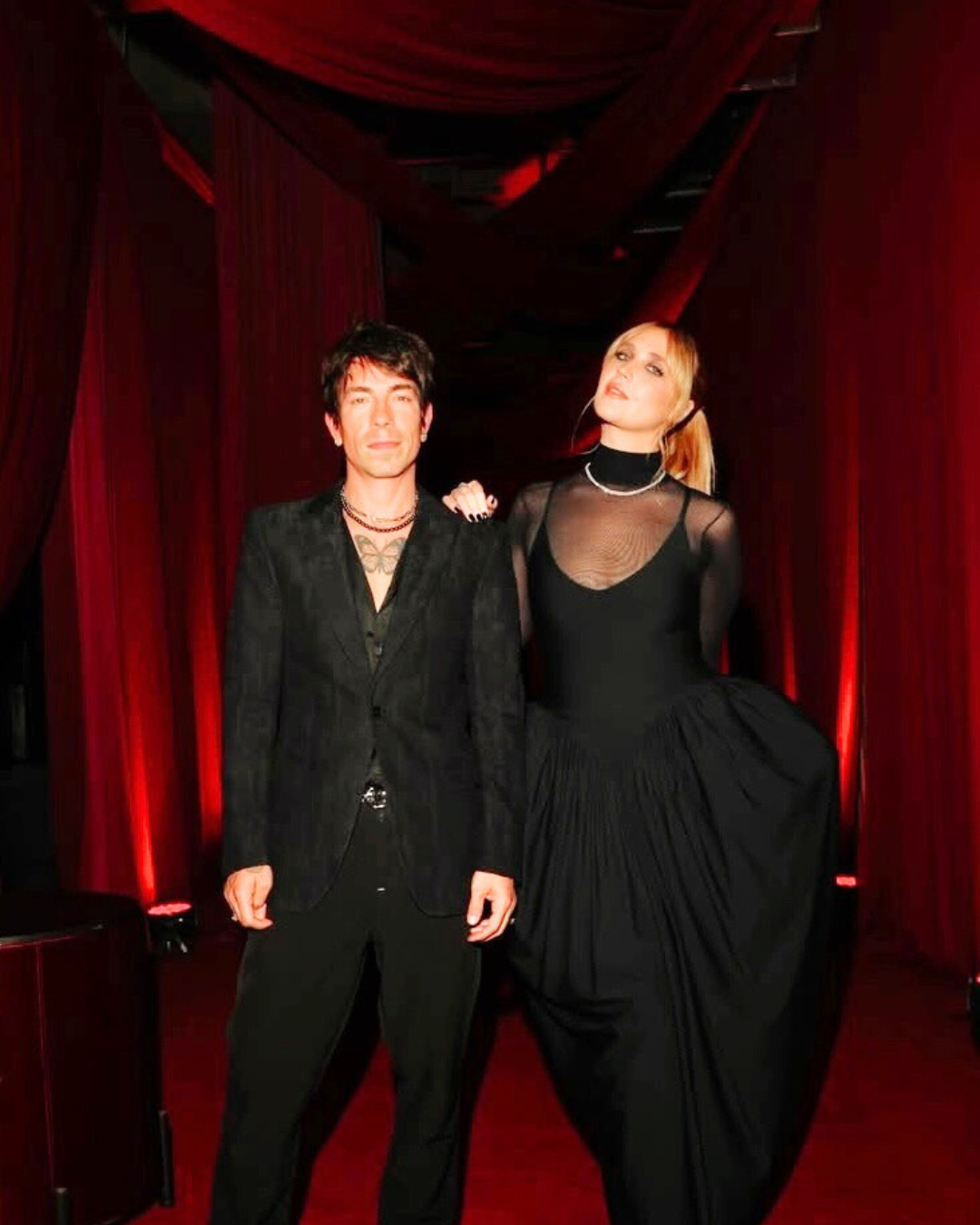 Homem e mulher posando juntos em ambiente com cortinas vermelhas, ela vestindo vestido preto com manga longa transparente, ele usando terno preto. Ela tem cabelo loiro e usa headset, ele tem cabelo escuro curto.