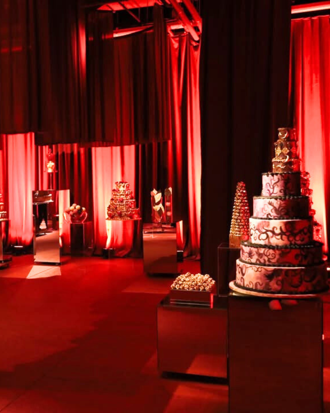 Salão decorado com cortinas vermelhas, várias festas de bolos requintados e decorações sofisticadas para uma celebração sofisticada e elegante.