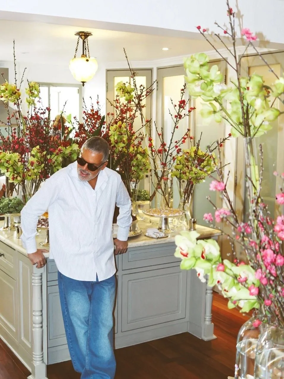 Homem com óculos escuros, camisa branca e jeans, em ambiente decorado com flores e vasos, perto de uma mesa de buffet.
