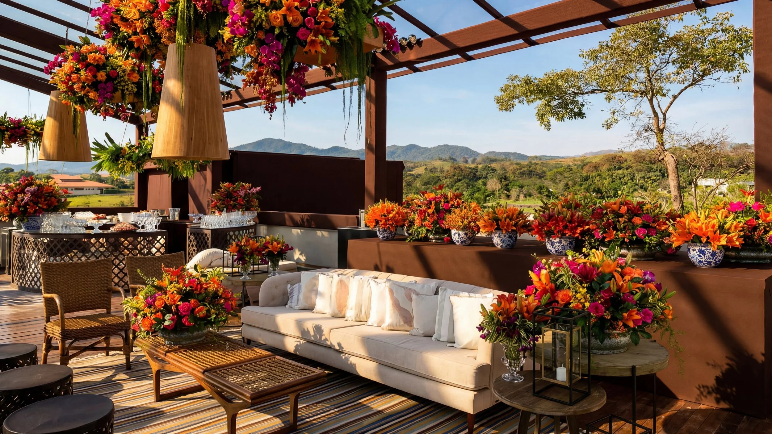 Deck de varanda decorada com flores coloridas em vasos, lounge de sofá com almofadas brancas, mesas de centro, cadeiras e iluminação suspensa, com vista para montanhas e árvores ao ar livre.